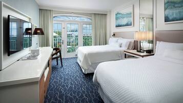 A hotel room with two beds, a desk with a flat-screen TV, a chair, and a balcony with a view.