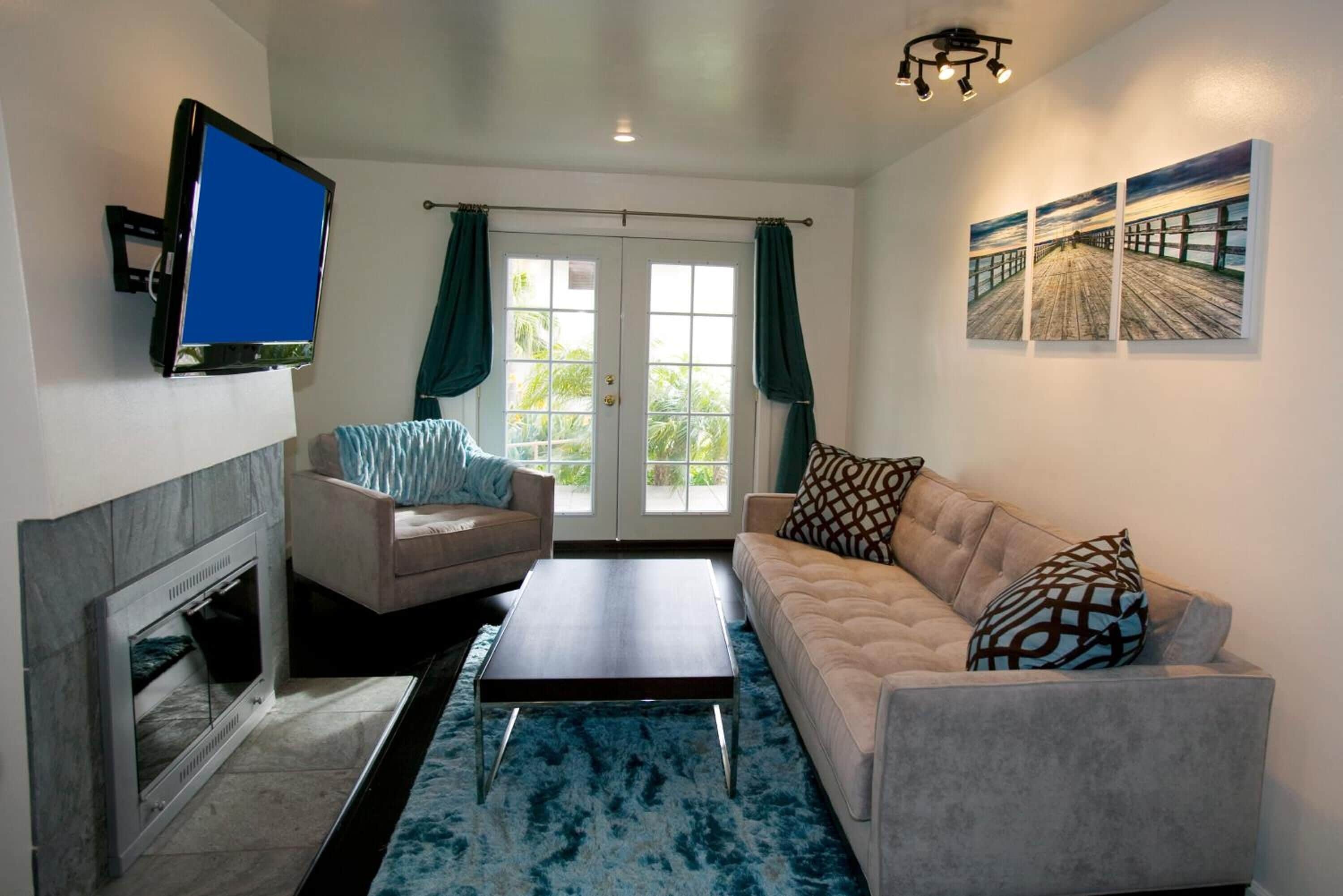 A living room with a fireplace, a flat-screen TV mounted on the wall, a sofa, a coffee table, and a large window with curtains.
