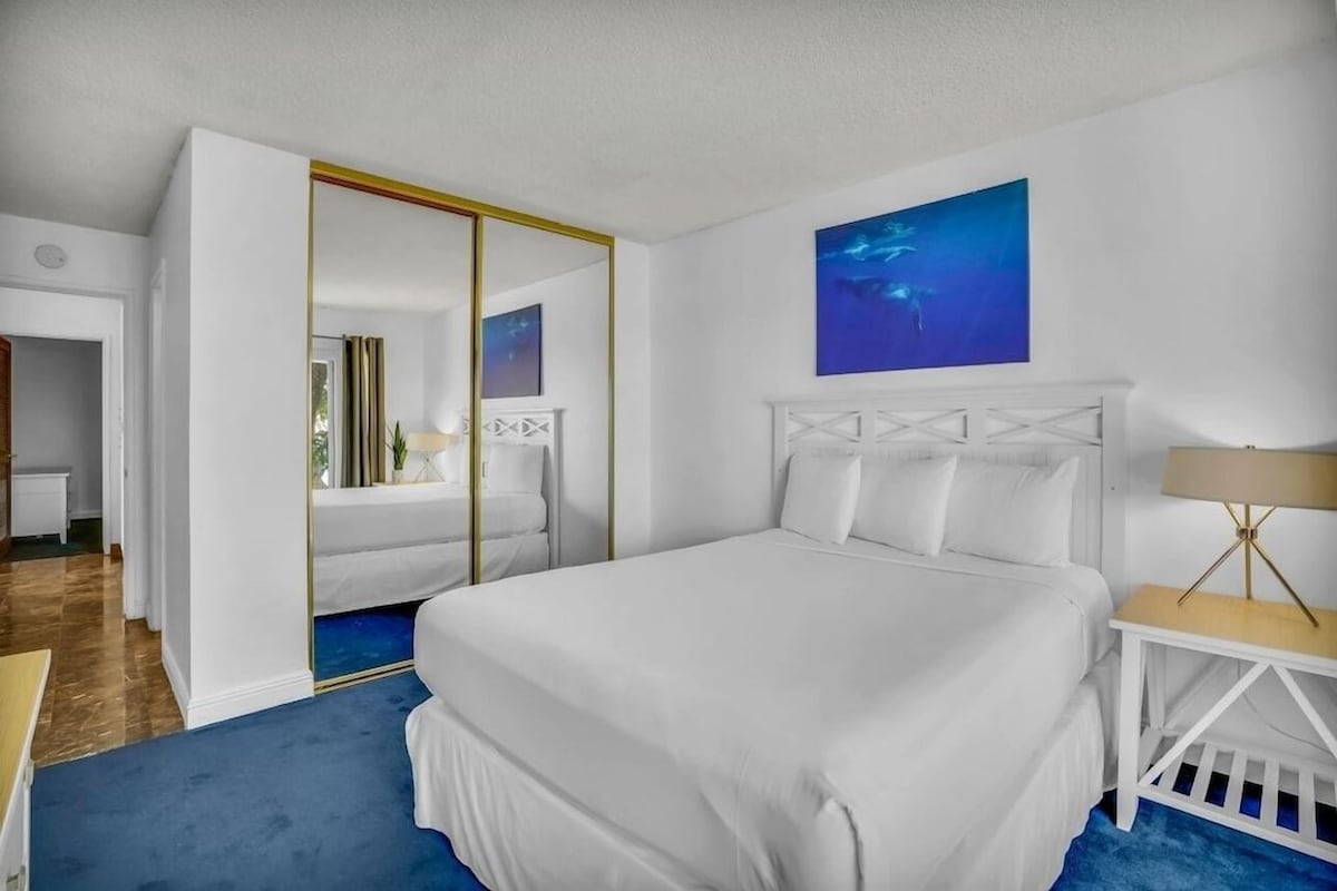 Habitación de hotel con una cama grande, cabecera blanca, mesita de noche con lámpara, alfombra azul y un cuadro en la pared.
