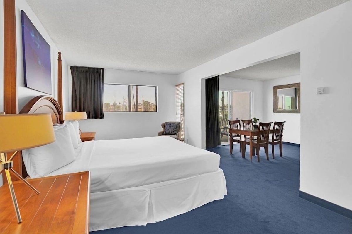 Habitación de hotel con cama, escritorio, silla, mesa de comedor con sillas y vista a la ciudad a través de la ventana.