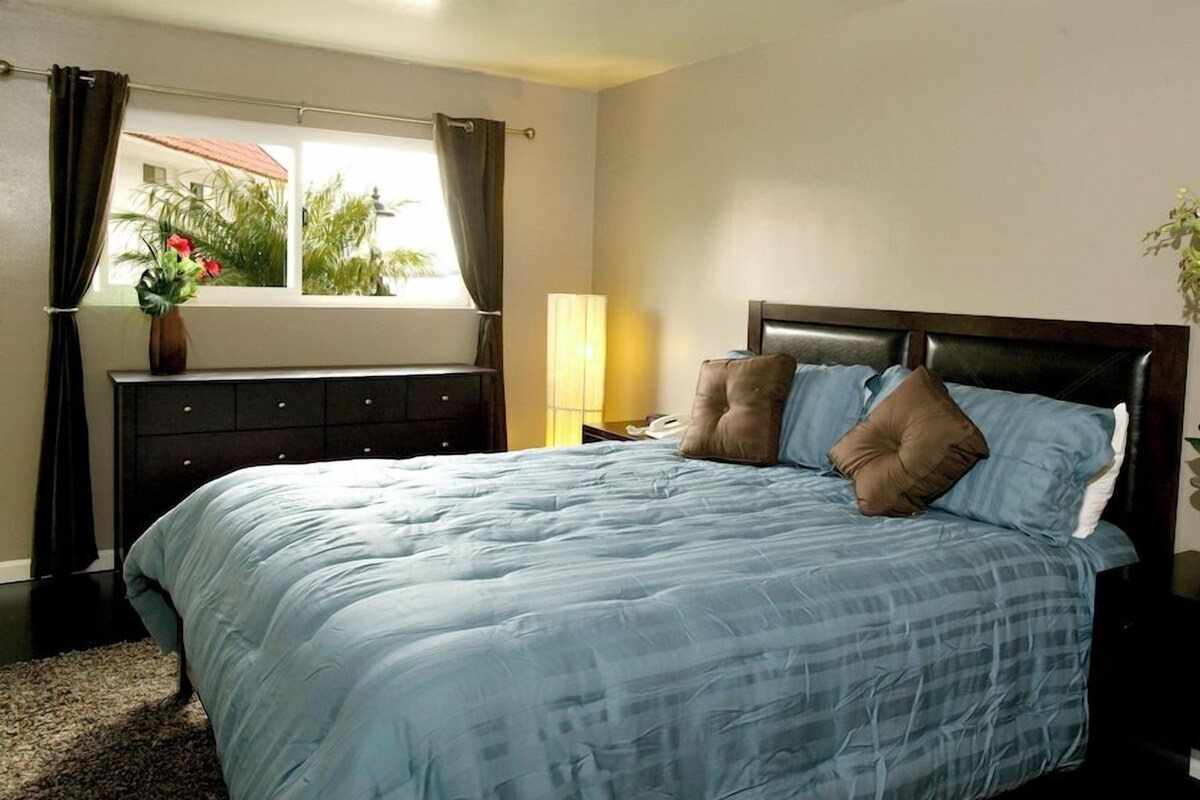Un dormitorio con cama, mesita de noche, lámpara, ventana con cortinas y una planta en maceta.