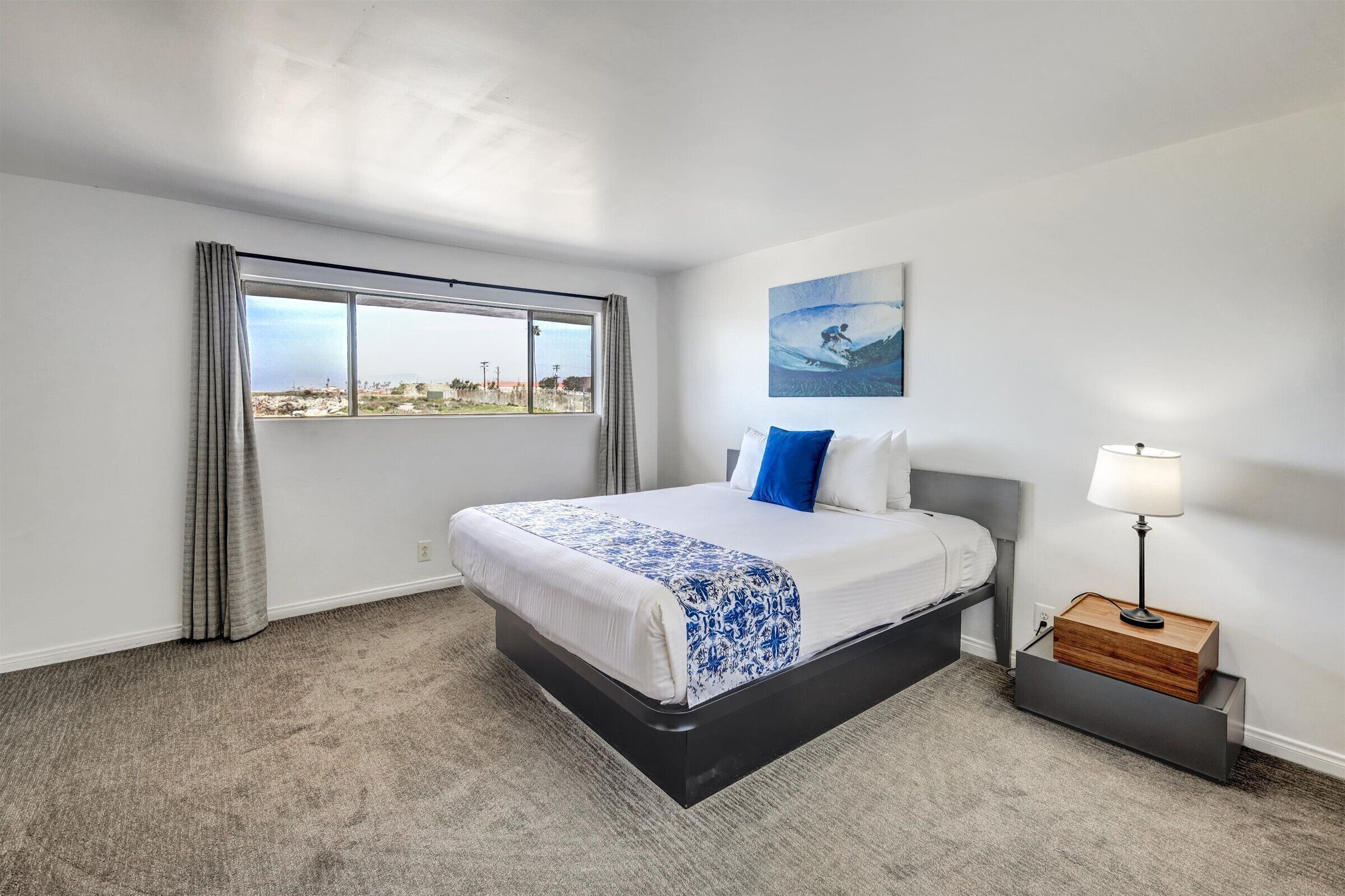 A hotel room with a bed, a nightstand, a lamp, a window with curtains, and a painting on the wall.