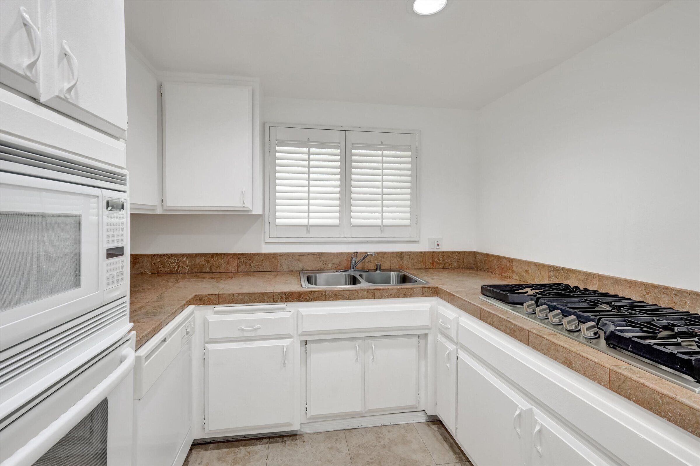 Una cocina con armarios blancos, un lavamanos doble, una estufa y una ventana con persianas.