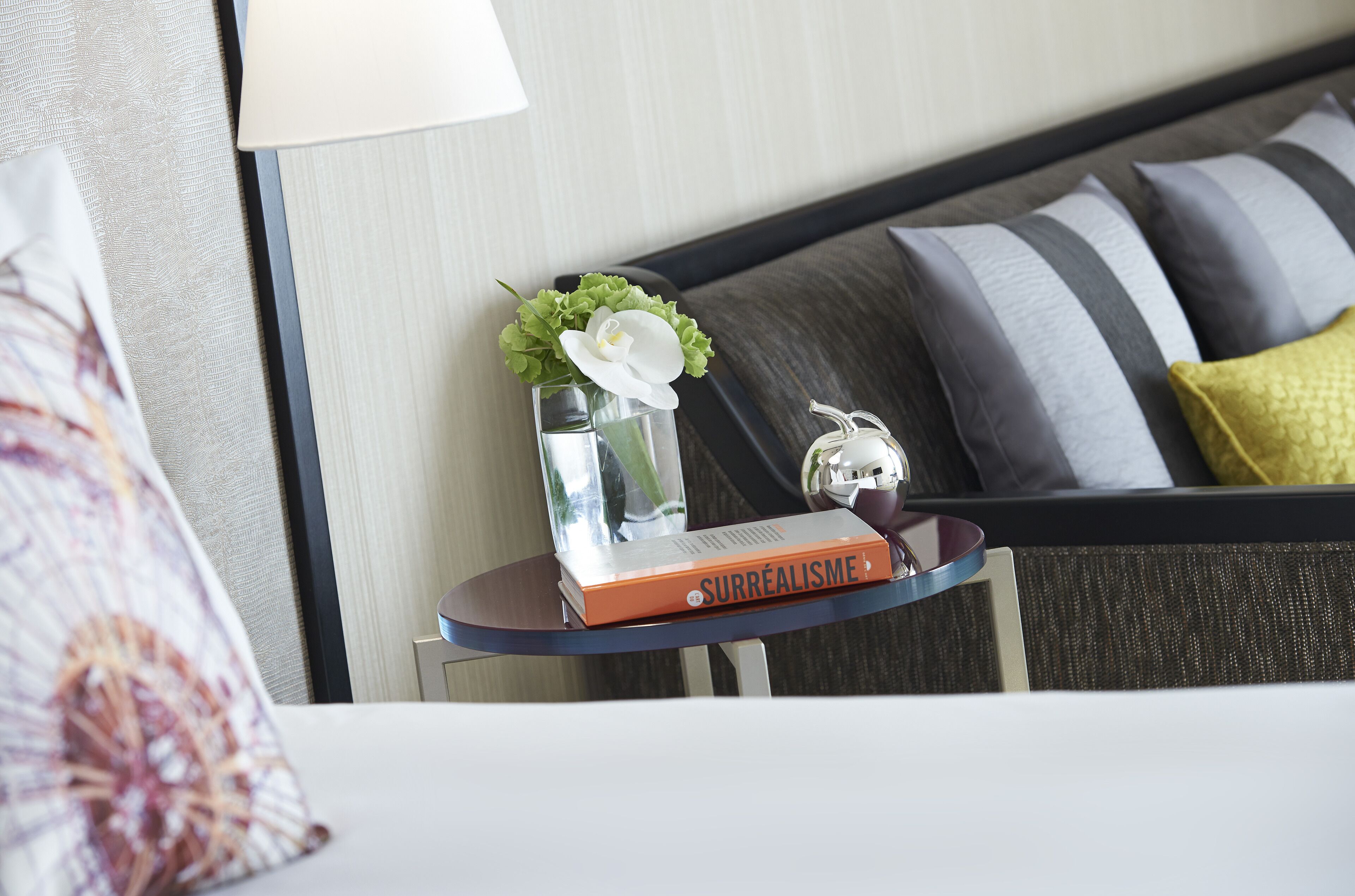 Une chambre d’hôtel bien rangée, avec un lit, une table de chevet, une lampe et un livre intitulé « SURRÉALISME ».