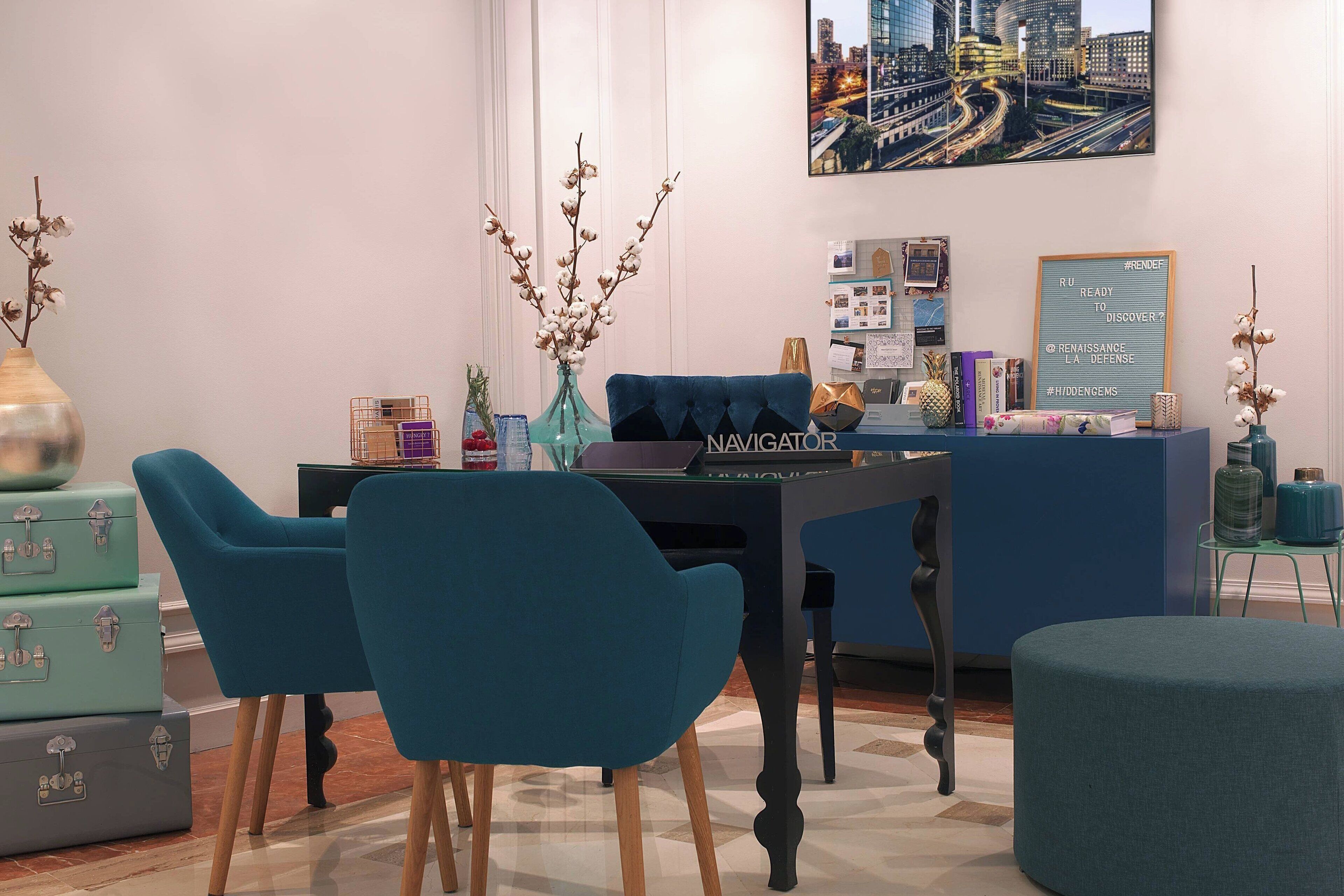 A modern dining area with a blue upholstered table and chairs, a vase with dried flowers, and a cityscape painting on the wall.