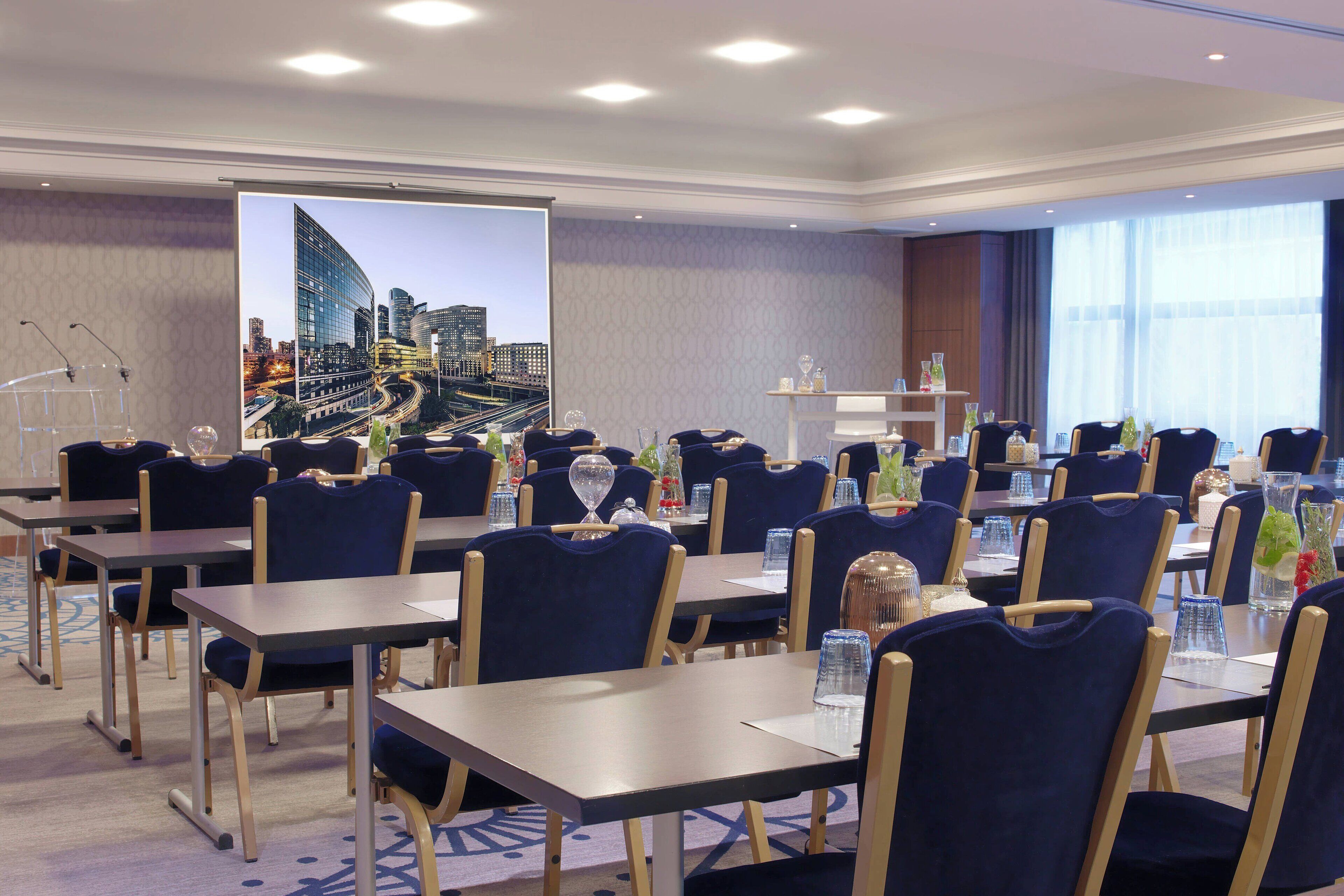 Un salón de conferencias con mesas y sillas, una pantalla de proyector y vistas a la ciudad.