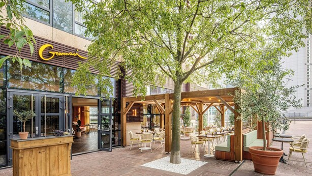 An outdoor cafe area with wooden seating, a large tree, and a building with a glass facade and a sign reading "Gramaï". - Renaissance Paris La Defense Hotel (Puteaux)