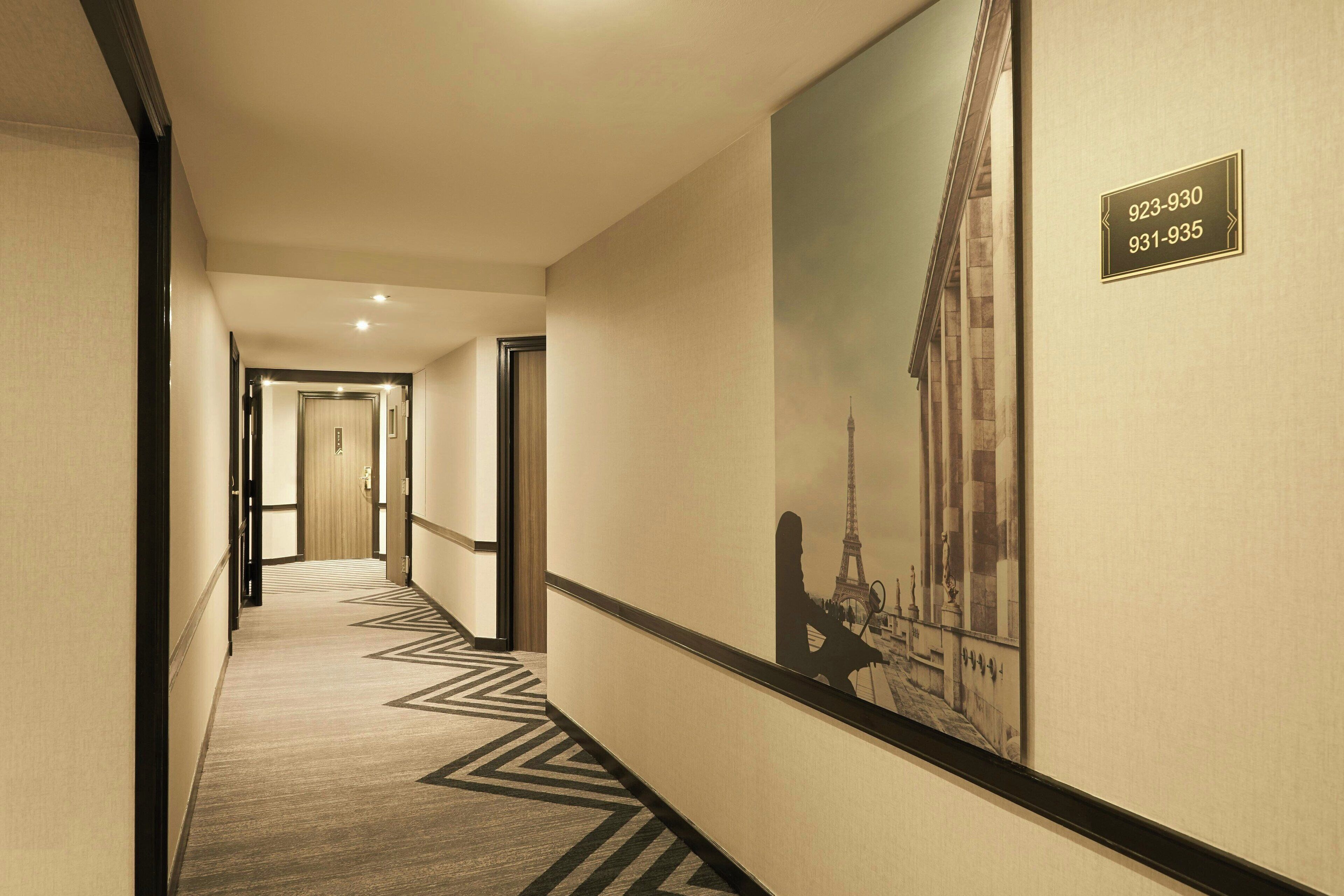 Passadís d'hotel amb una catifes estampada, un quadre gran amb una vista de la ciutat i un cartell amb números de telèfon.