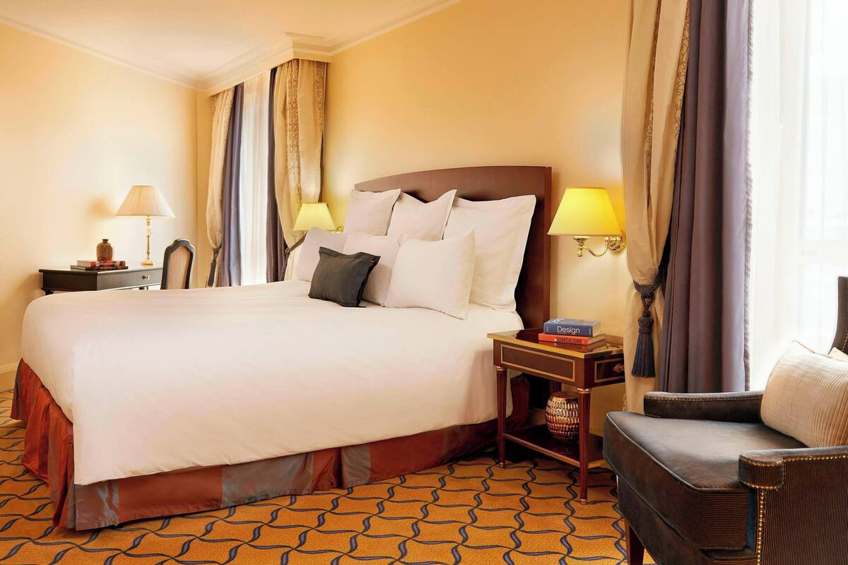 Quarto de hotel com uma cama grande, duas mesas de cabeceira com luminárias, uma cadeira e um tapete estampado.