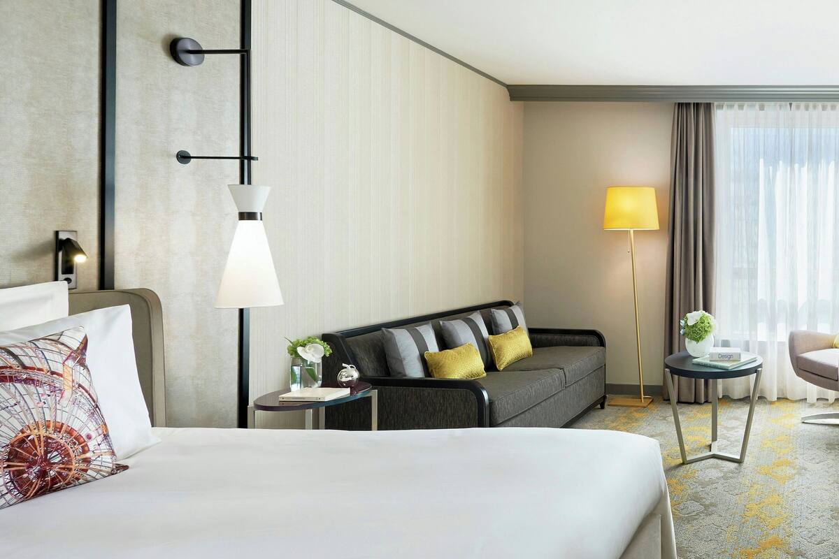 Quarto de hotel com uma cama grande, um sofá, uma pequena mesa com abajur e uma cadeira.