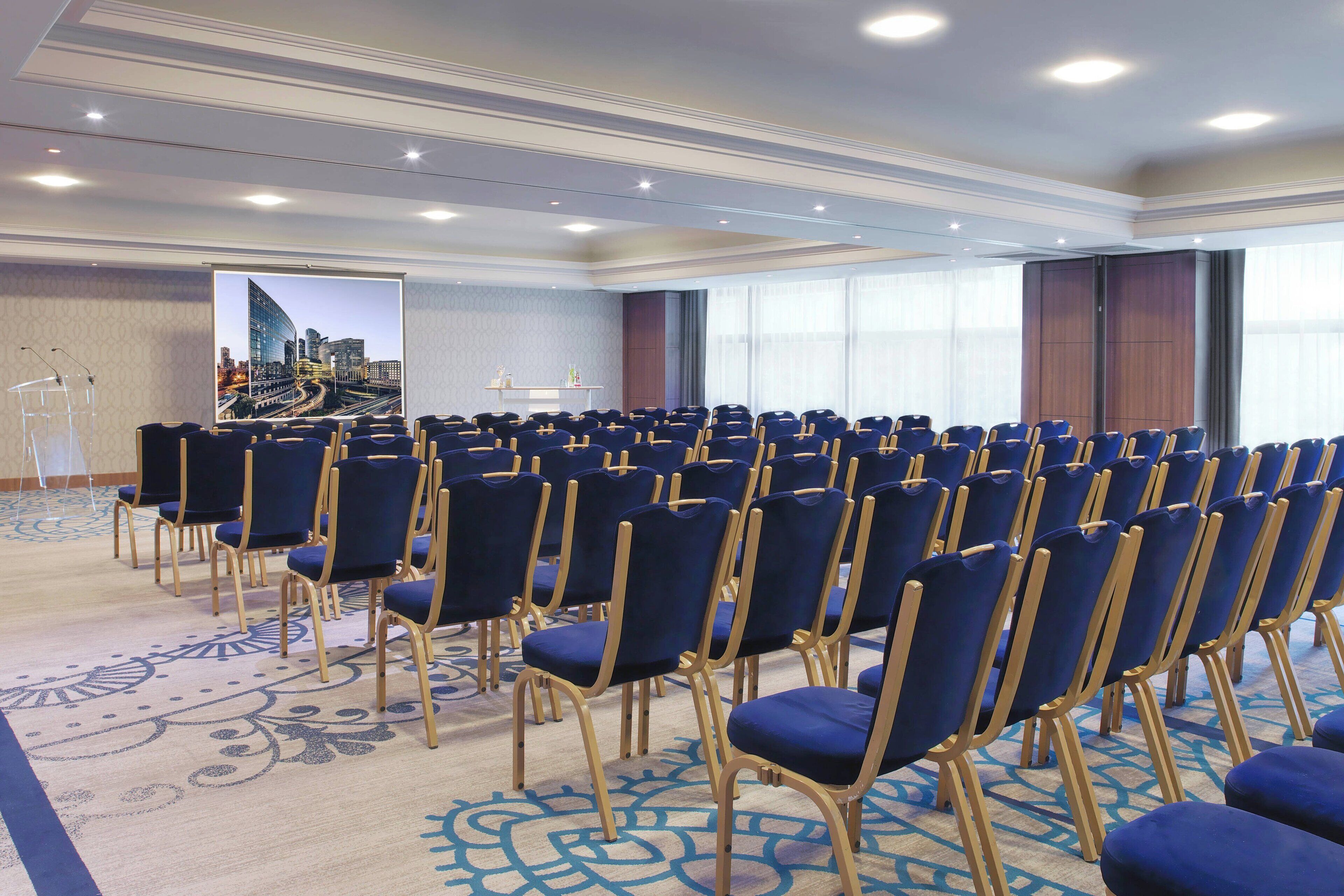 A conference room with rows of blue chairs, a podium, and a large screen displaying a cityscape.