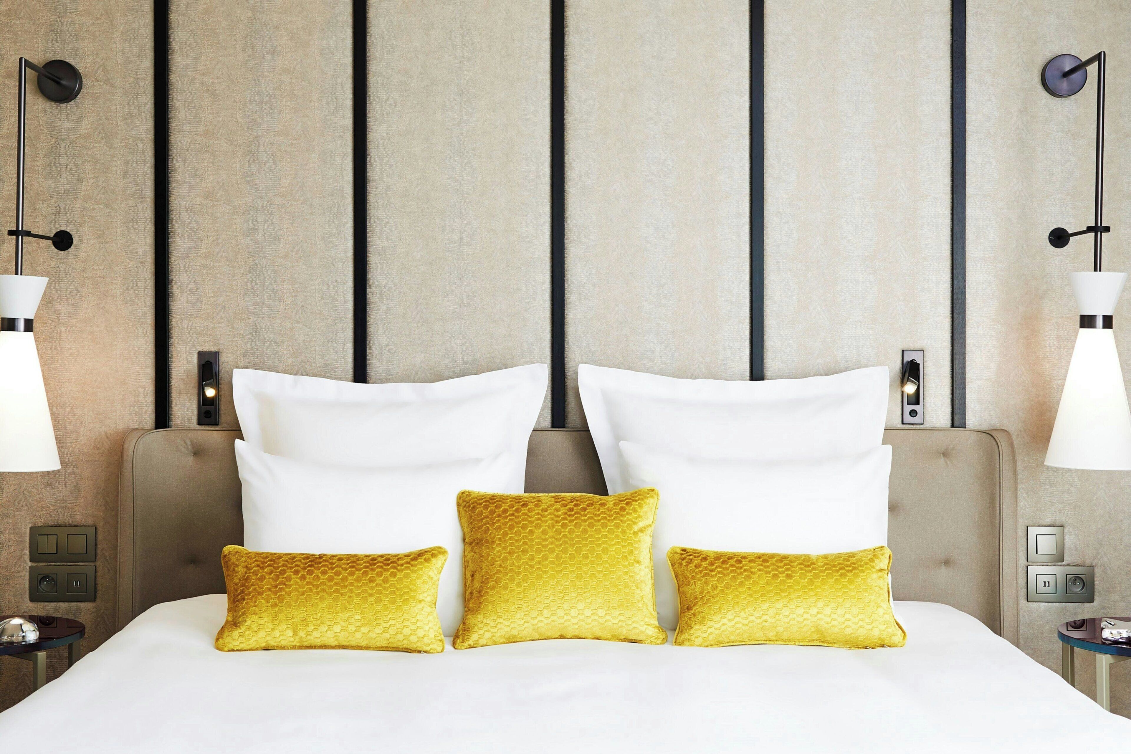 Habitación de hotel con una cama grande, tres almohadas blancas y tres almohadas doradas. Hay dos mesitas de noche con lámparas y un panel de control en la pared.