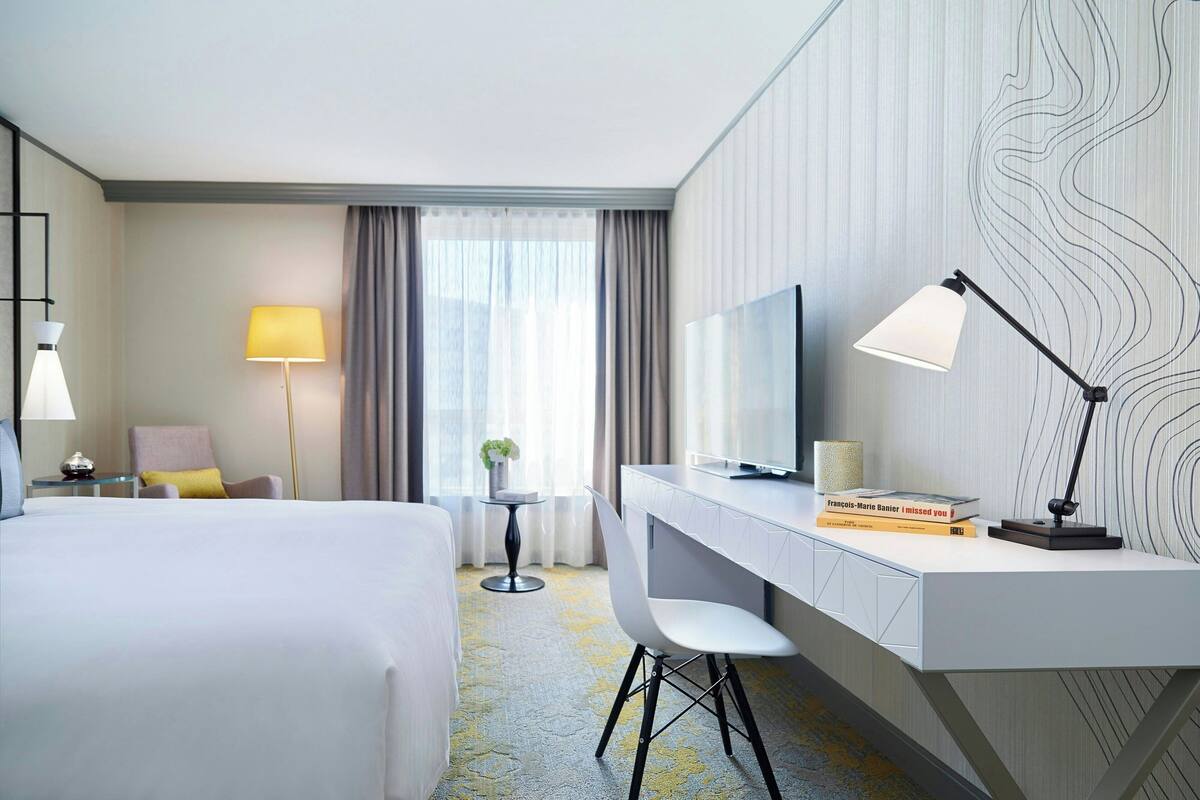 Quarto de hotel com cama, escrivaninha, cadeira, televisão e abajur.