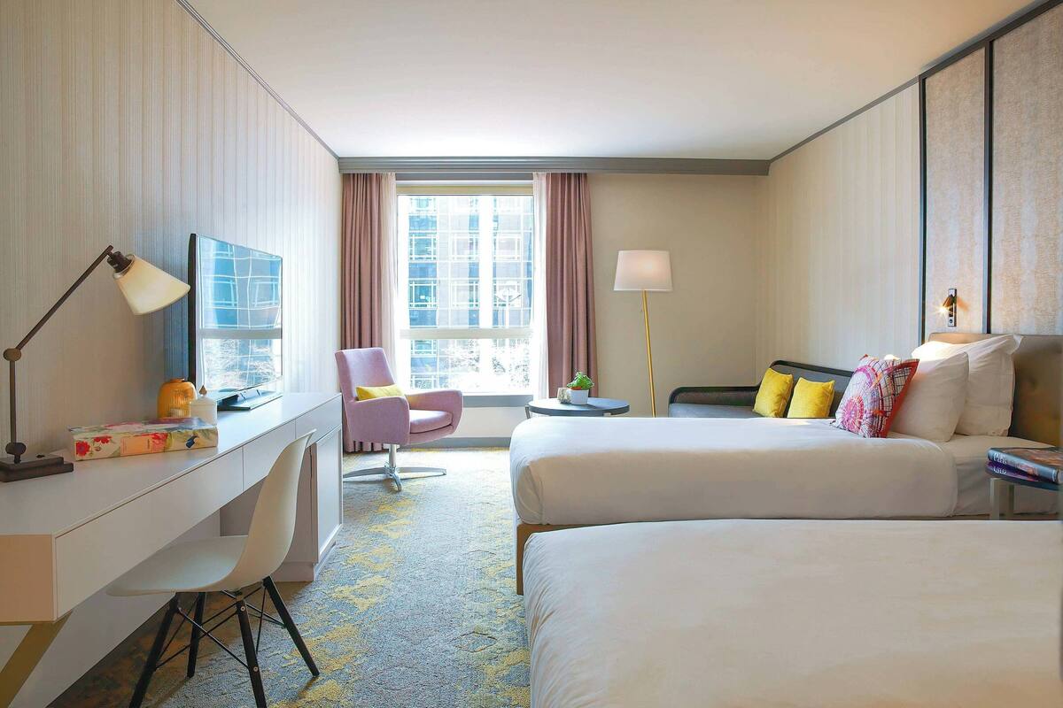 Quarto de hotel com escrivaninha, duas camas, uma cadeira, abajur e janela com cortinas.
