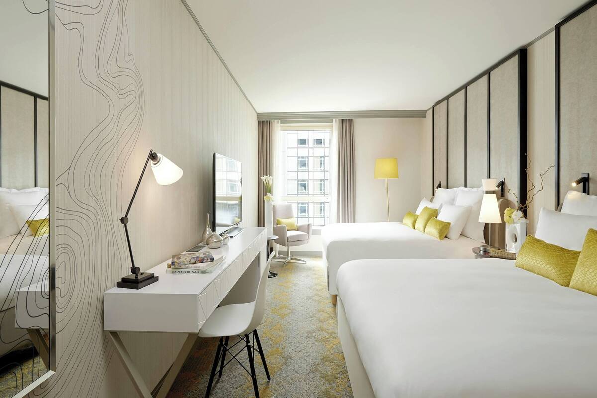 Quarto de hotel com escrivaninha, duas camas, uma cadeira e uma janela grande.