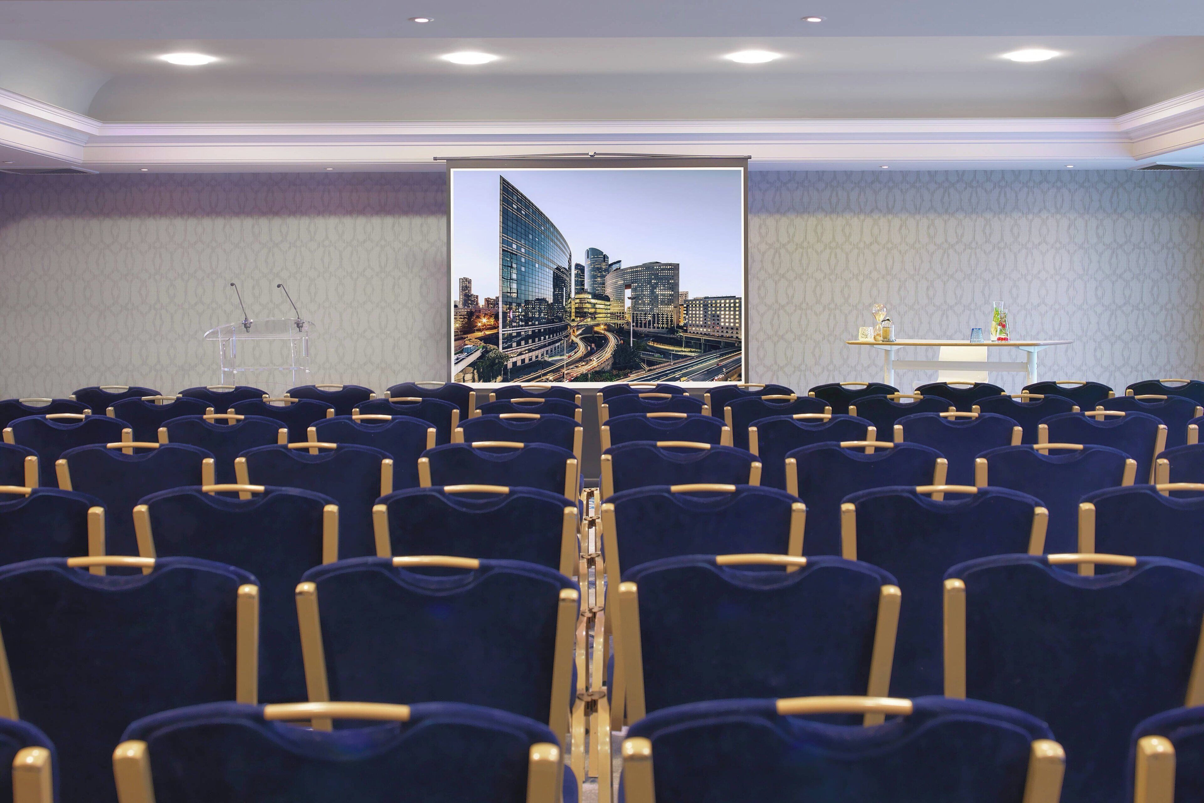 A conference room with rows of blue chairs, a podium, and a large cityscape view on the screen.