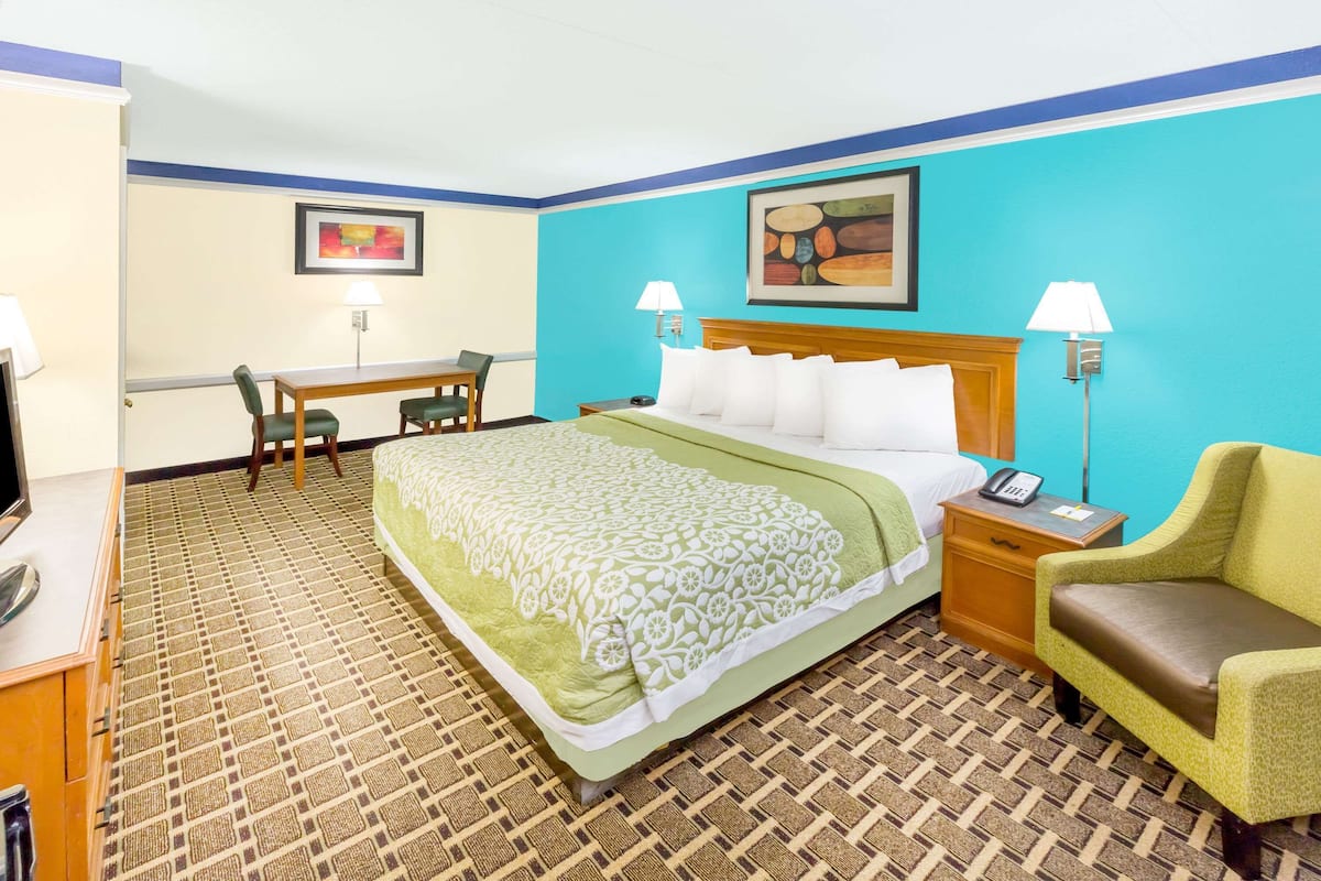 A hotel room with a bed, bedside table, lamp, chair, desk, and a painting on the wall.