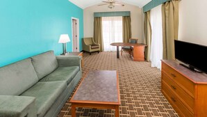 Ein Hotelzimmer mit einem grünen Sofa, einem Holztisch, einem Fernseher auf einer Holzkommode, einem Deckenventilator und einem Fenster mit Vorhängen.