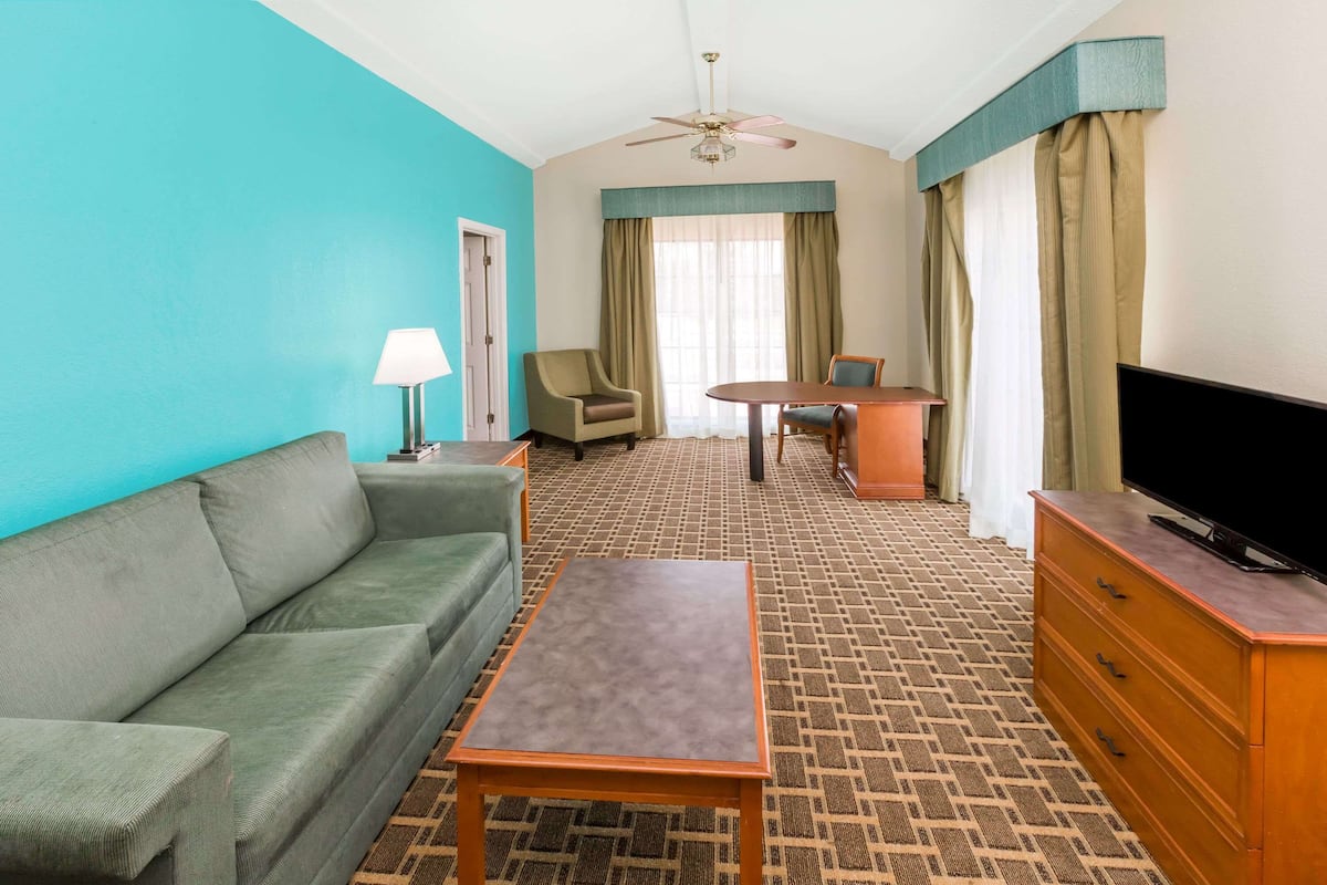 A hotel room with a green sofa, a wooden coffee table, a TV on a wooden dresser, a ceiling fan, and a window with curtains.