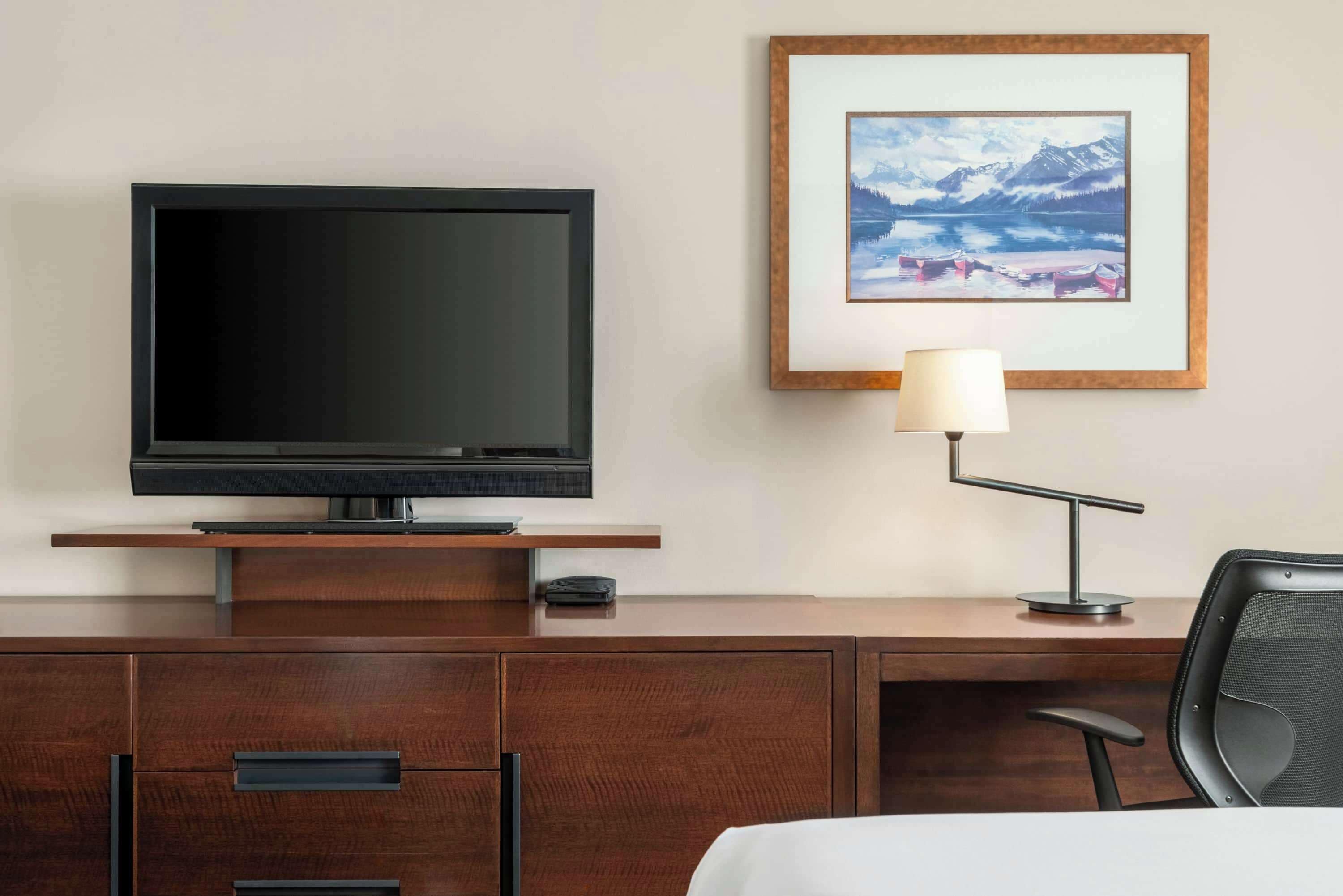 A hotel room with a flat-screen TV on a wooden stand, a desk with a lamp, and a framed picture of a mountain landscape.