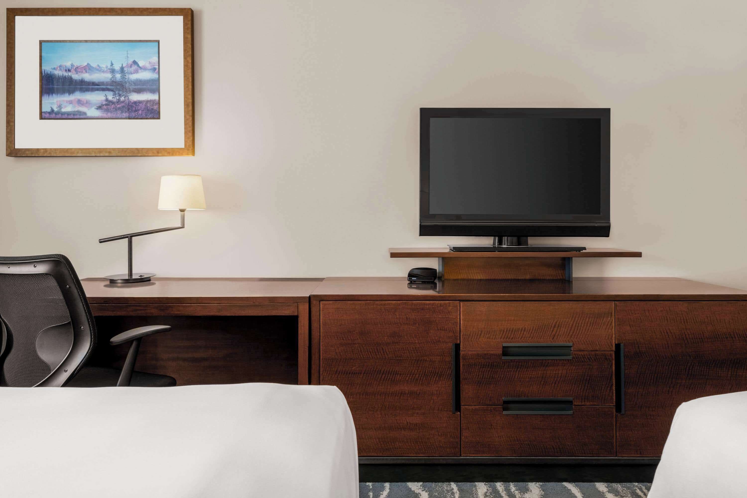 Quarto de hotel com escrivaninha de madeira, televisão, luminária, cadeira e cama.