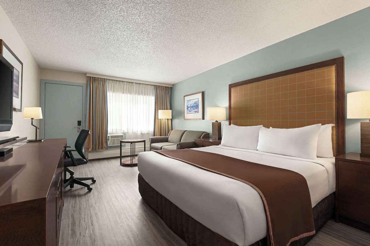 A hotel room with a large bed, a desk, two chairs, a TV, and a window with curtains.