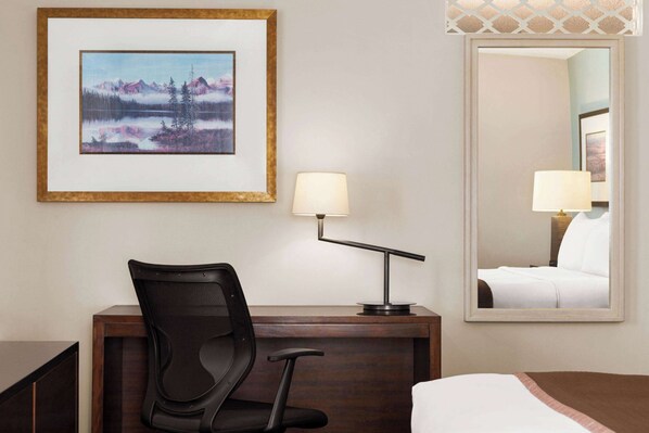 A hotel room with a desk, chair, lamp, bed, and a framed picture of a mountain landscape.