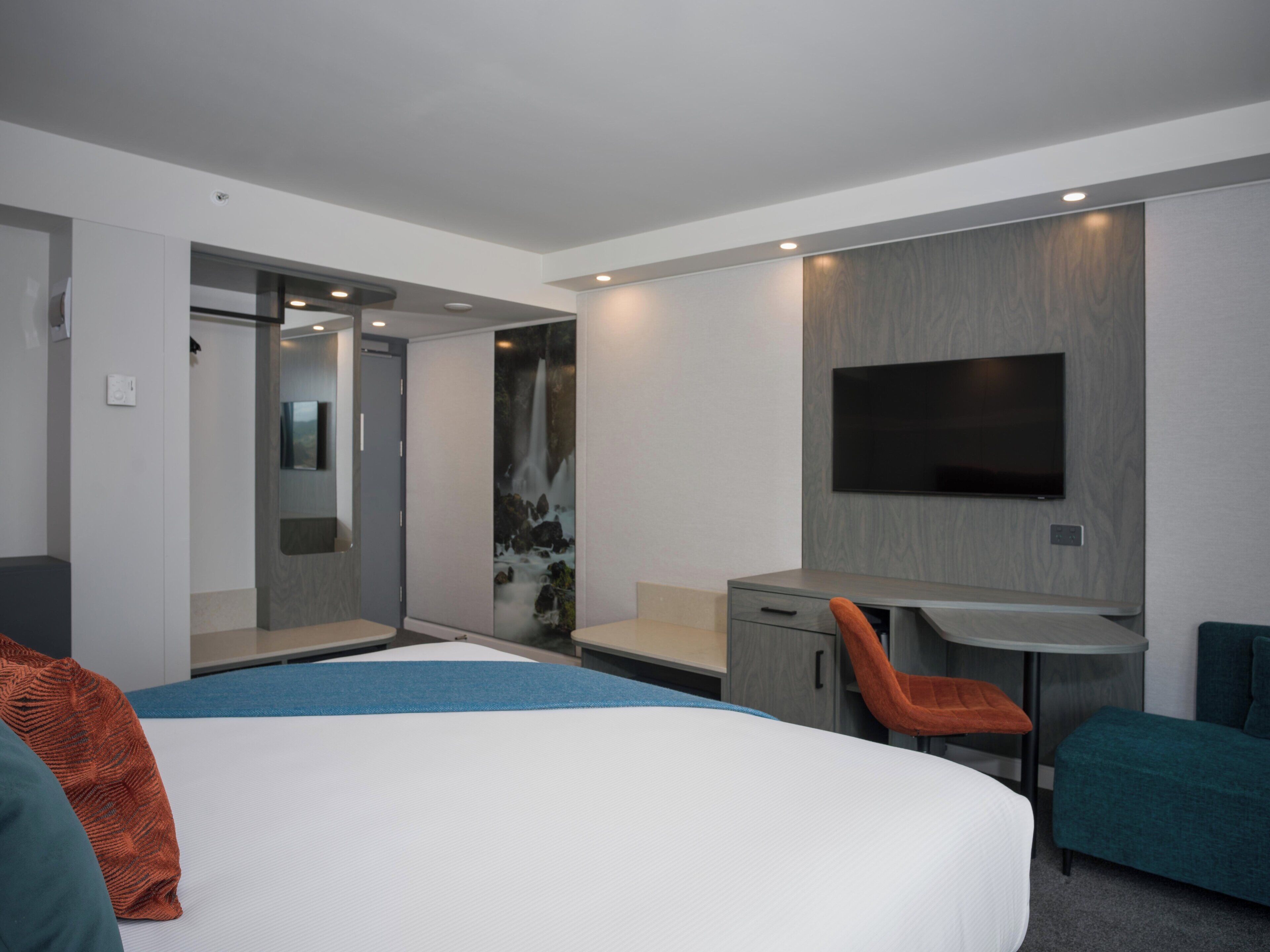Moderni hotellihuone, jossa on suuri sänky, työpöytä tuolilla, seinälle asennettu taulutelevisio ja suihkutila.