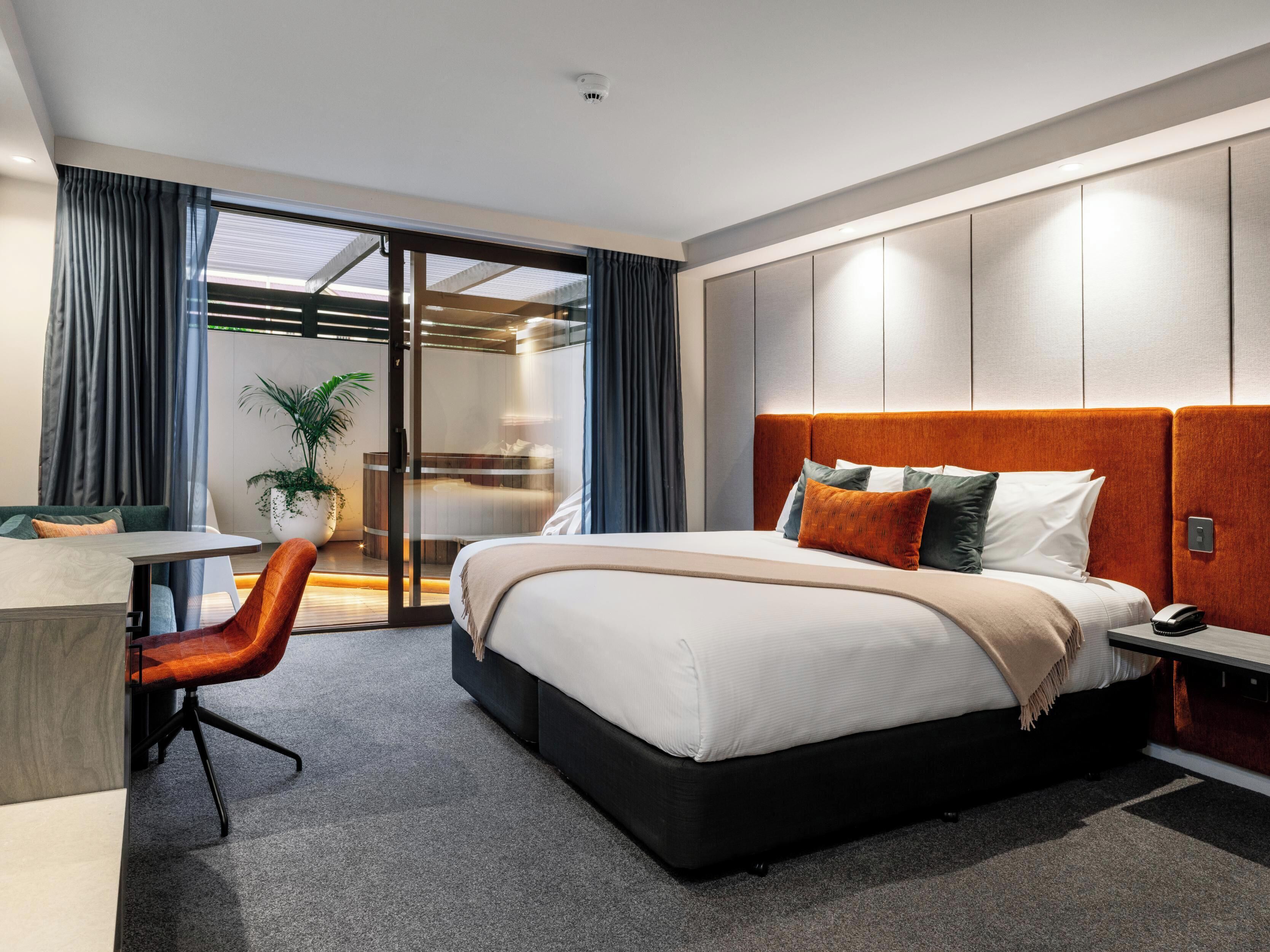 A modern hotel room with a large bed, a desk, a chair, and a sliding glass door leading to a balcony.