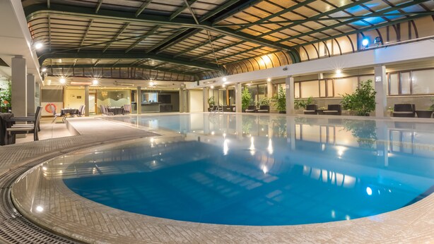 An indoor swimming pool with a curved edge, surrounded by lounge chairs and tables, under a high ceiling with a grid pattern.