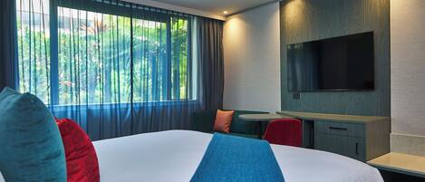 A hotel room with a large bed, a desk, a TV, and a view of greenery through the window.