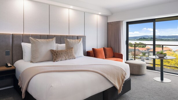 A modern bedroom with a large bed, a nightstand, a seating area, and a view of a landscape through large windows. - Millennium Hotel Rotorua (Rotorua)