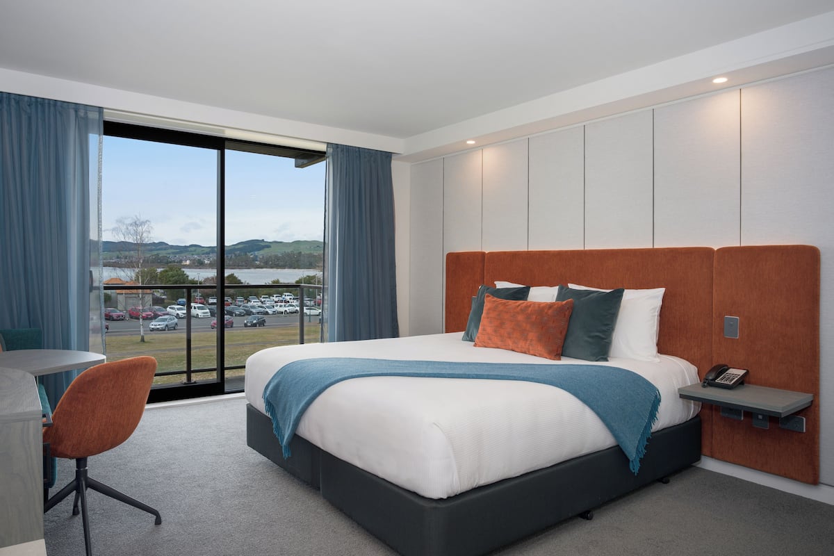 A modern hotel room with a large bed, a desk, and a view of a parking lot and hills.