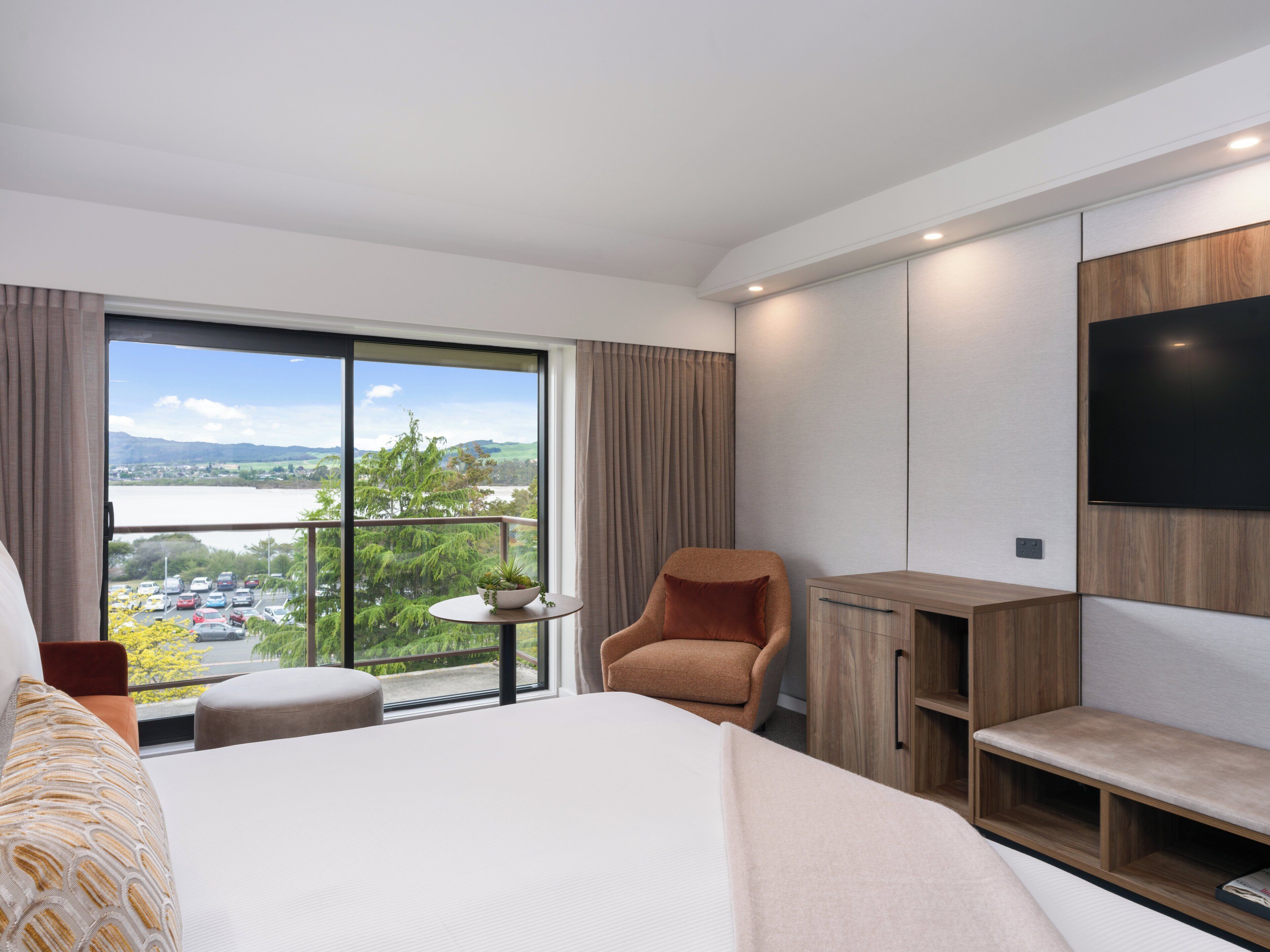 A modern hotel room with a large bed, a seating area with a chair and a small table, a TV mounted on the wall, and a balcony with a view of the landscape.