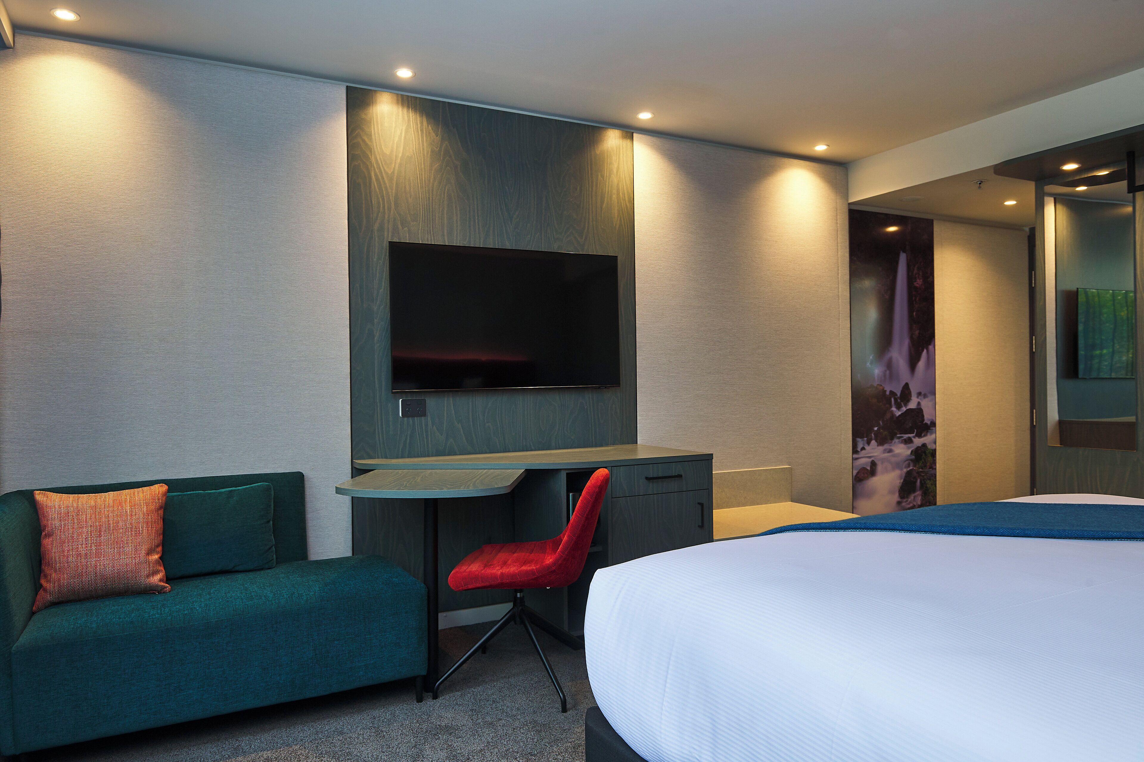 A modern hotel room with a bed, a desk, a red chair, and a TV mounted on the wall.