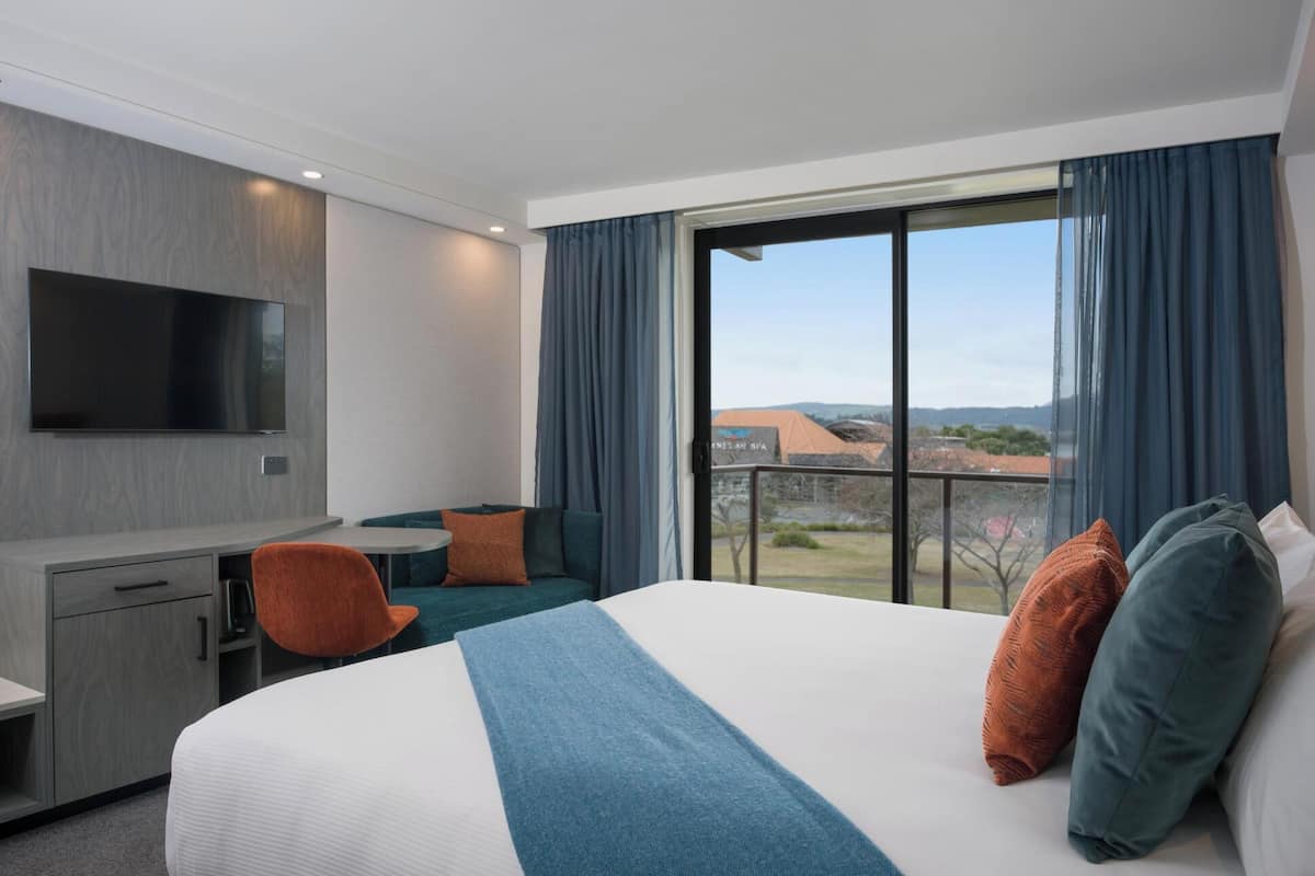 A hotel room with a bed, a desk, a chair, a TV, and a large window with a view of buildings and hills.
