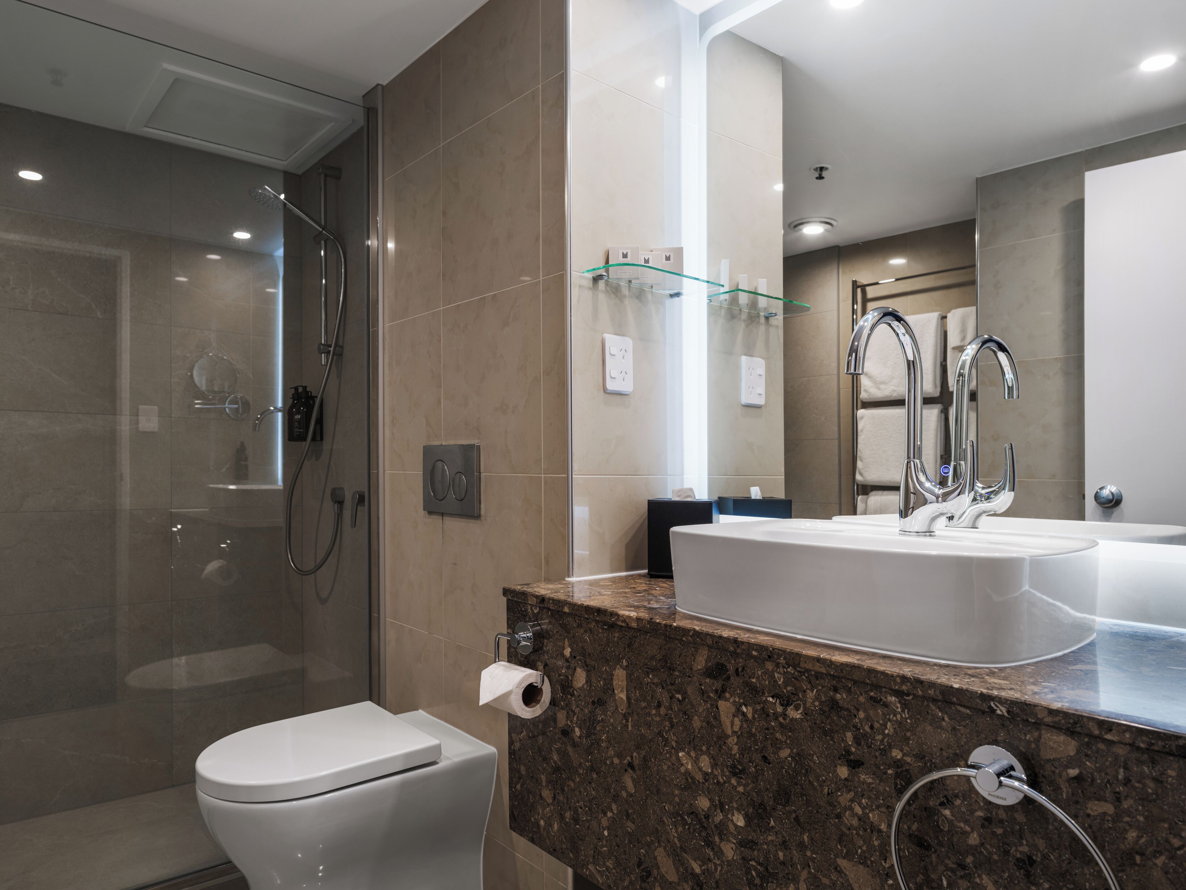 A modern bathroom with a glass-enclosed shower, a white toilet, a marble countertop, a large sink with a mirror, and a towel rack.