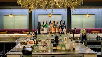 Uma área de bar com diversas garrafas de bebidas, copos e uma planta pendente decorativa.