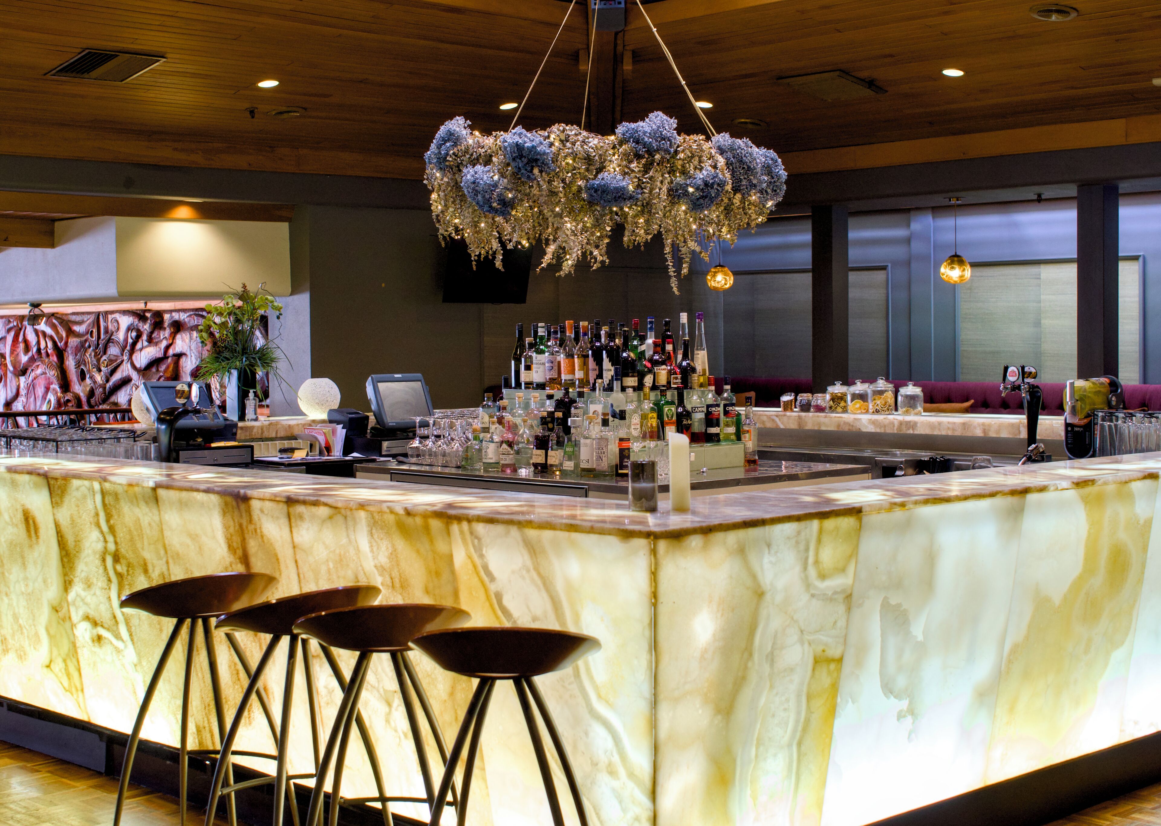 A bar area with a marble countertop, stools, a variety of bottles, and a decorative hanging light fixture.