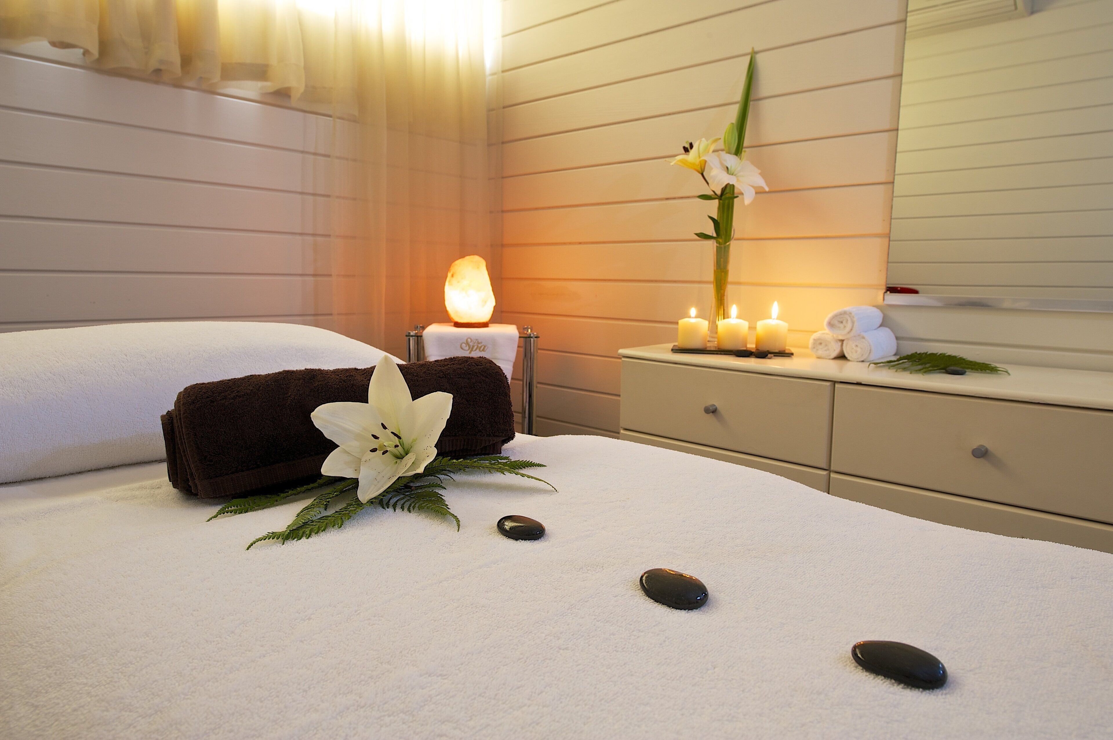 A spa room with a massage table, towels, a heated stone, candles, and a flower arrangement.