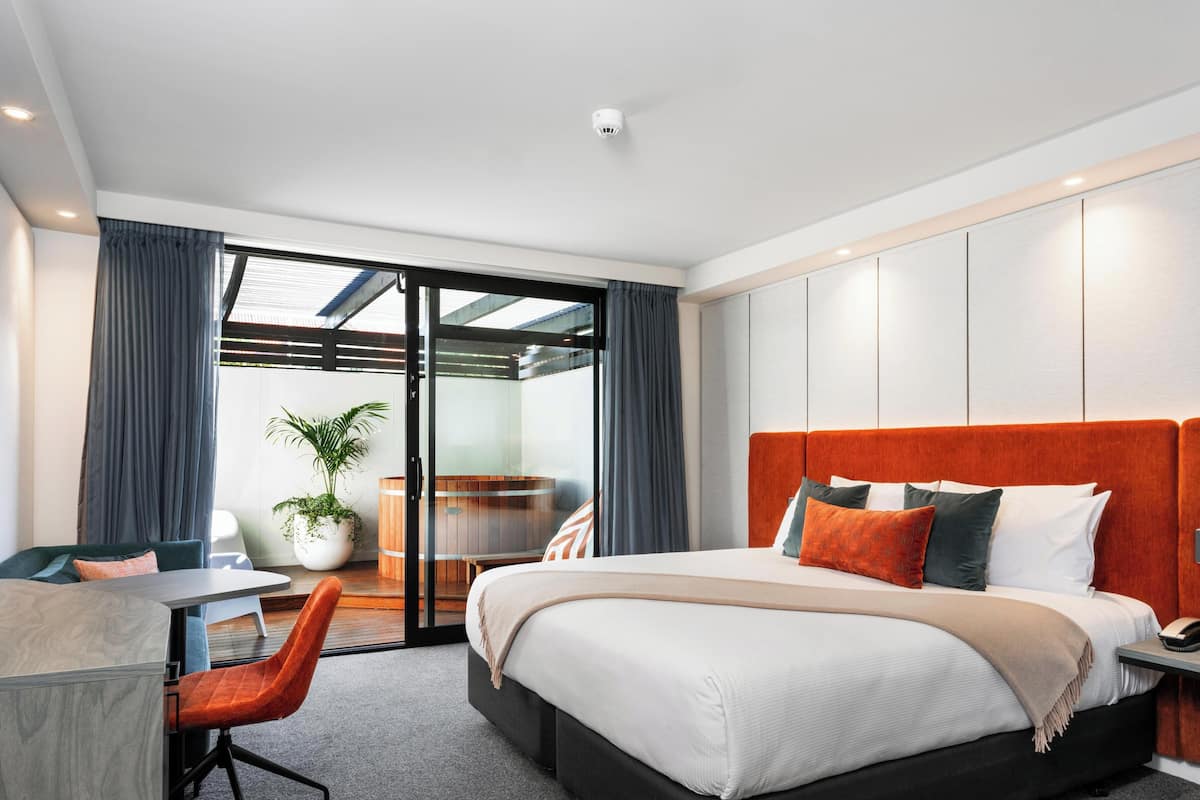 A modern hotel room with a large bed, a desk, and a chair. There is a sliding glass door leading to a balcony with a view.