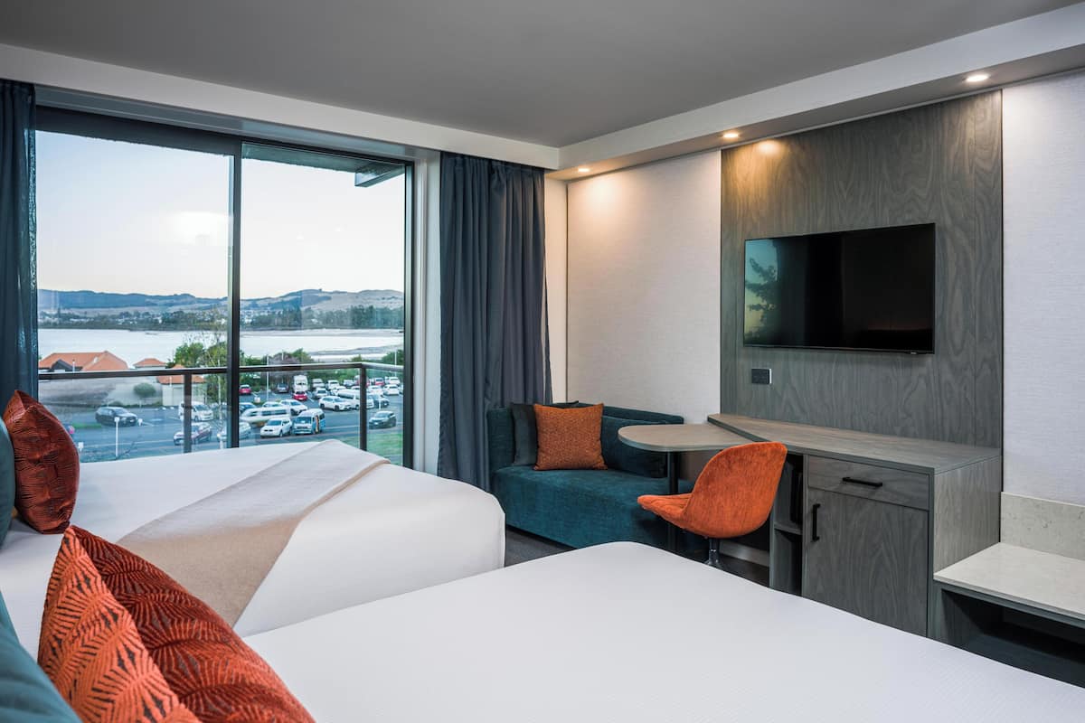 A hotel room with two beds, a desk, a TV, and a view of a parking lot and landscape.