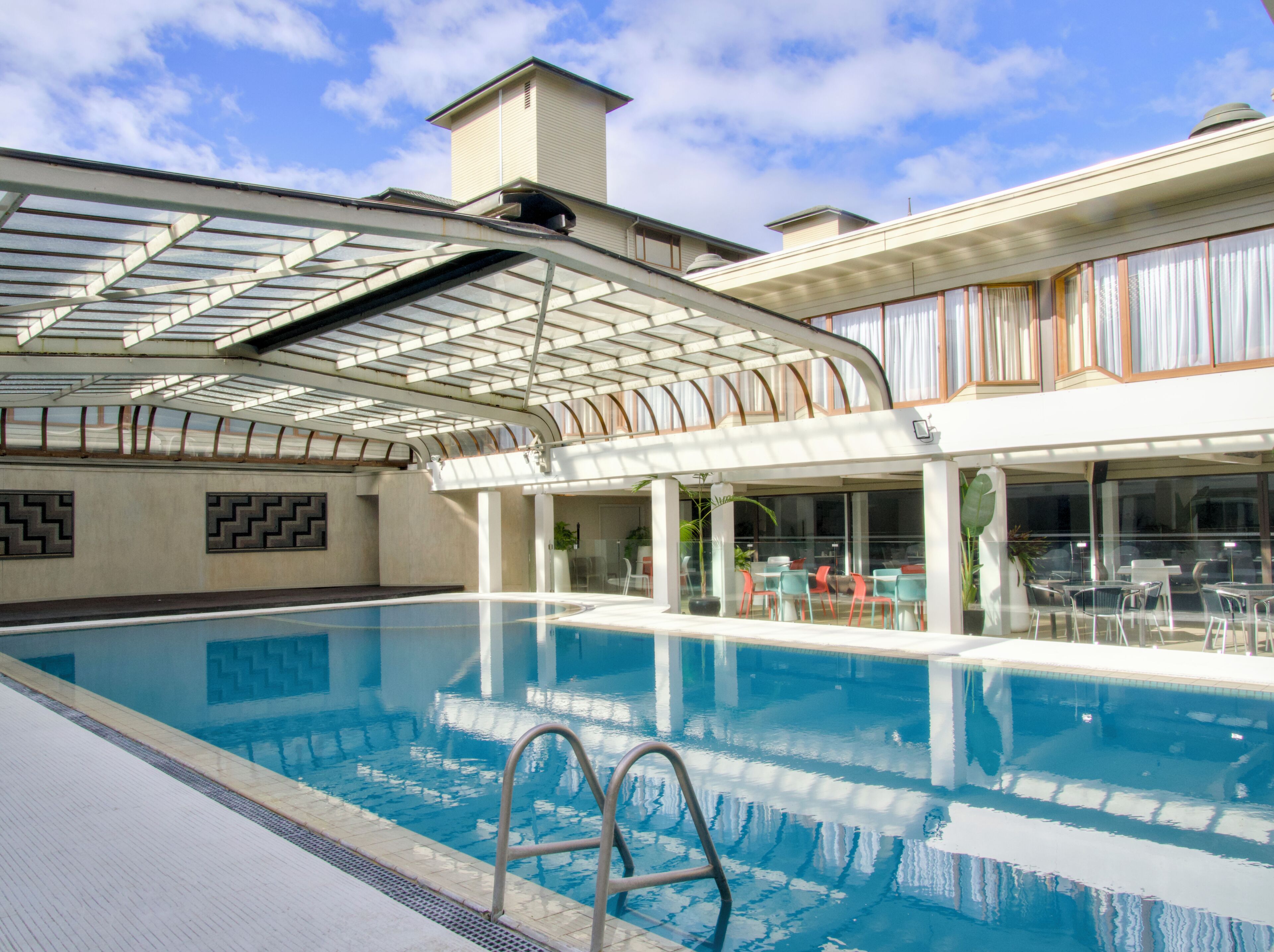 Una piscina con cerramiento de vidrio, mobiliario exterior y un edificio con grandes ventanales.