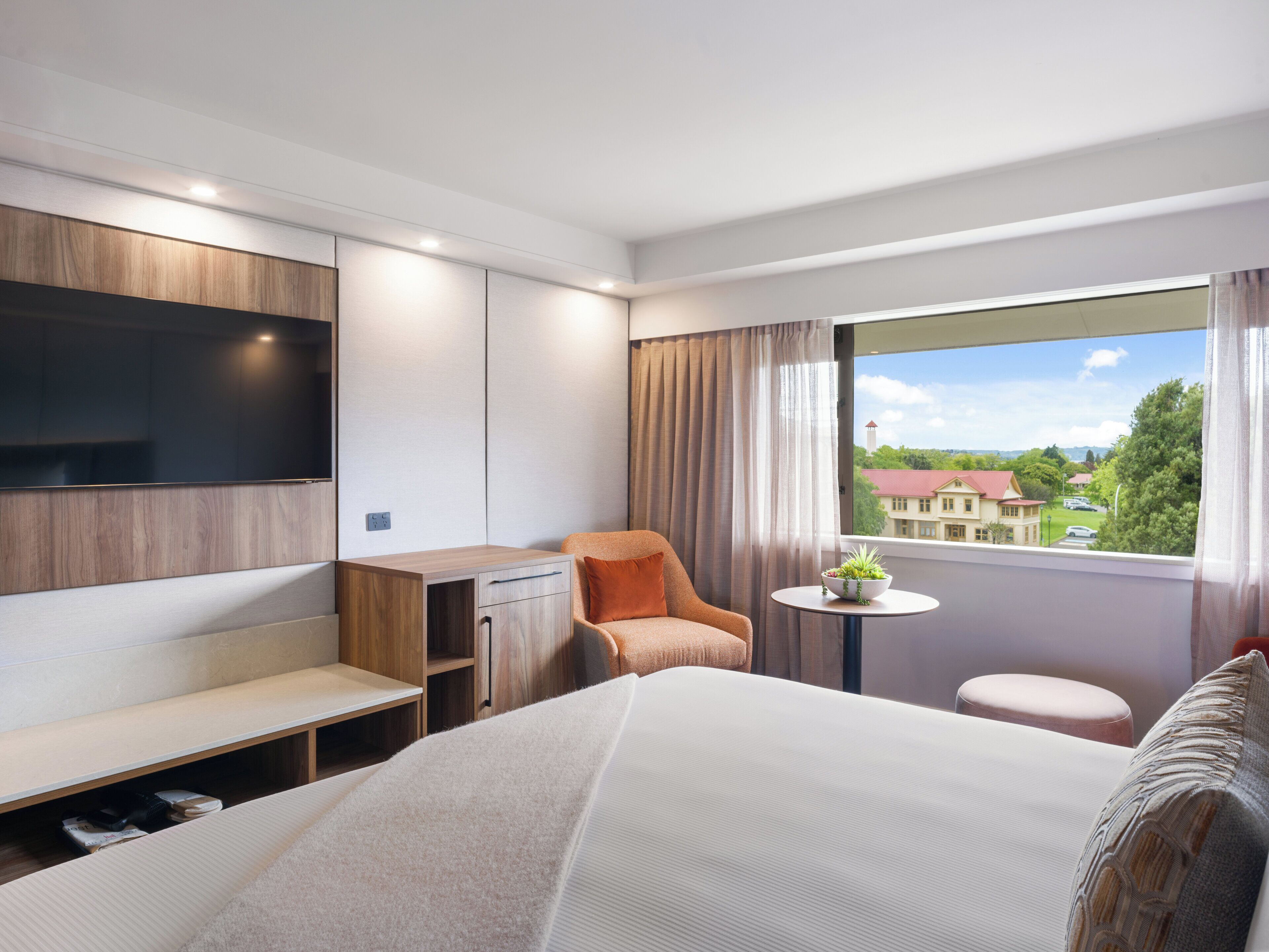 A modern hotel room with a large bed, a desk, a chair, a small table with a plant, and a TV mounted on the wall.
