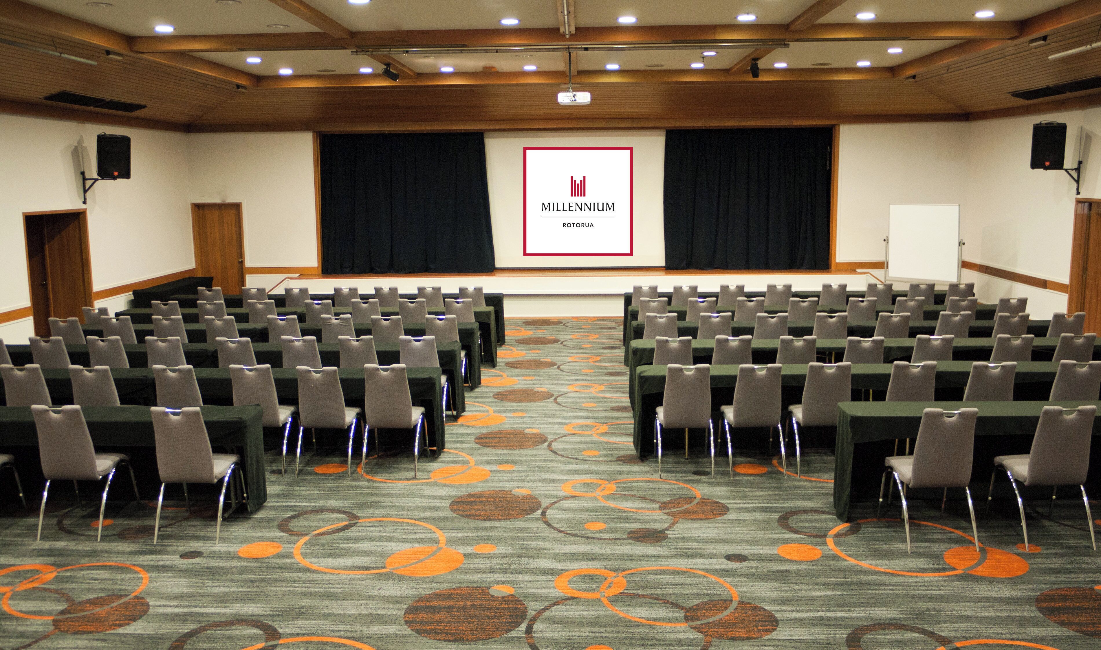 A conference room with rows of chairs, a stage with a screen, and a carpet with circular patterns.