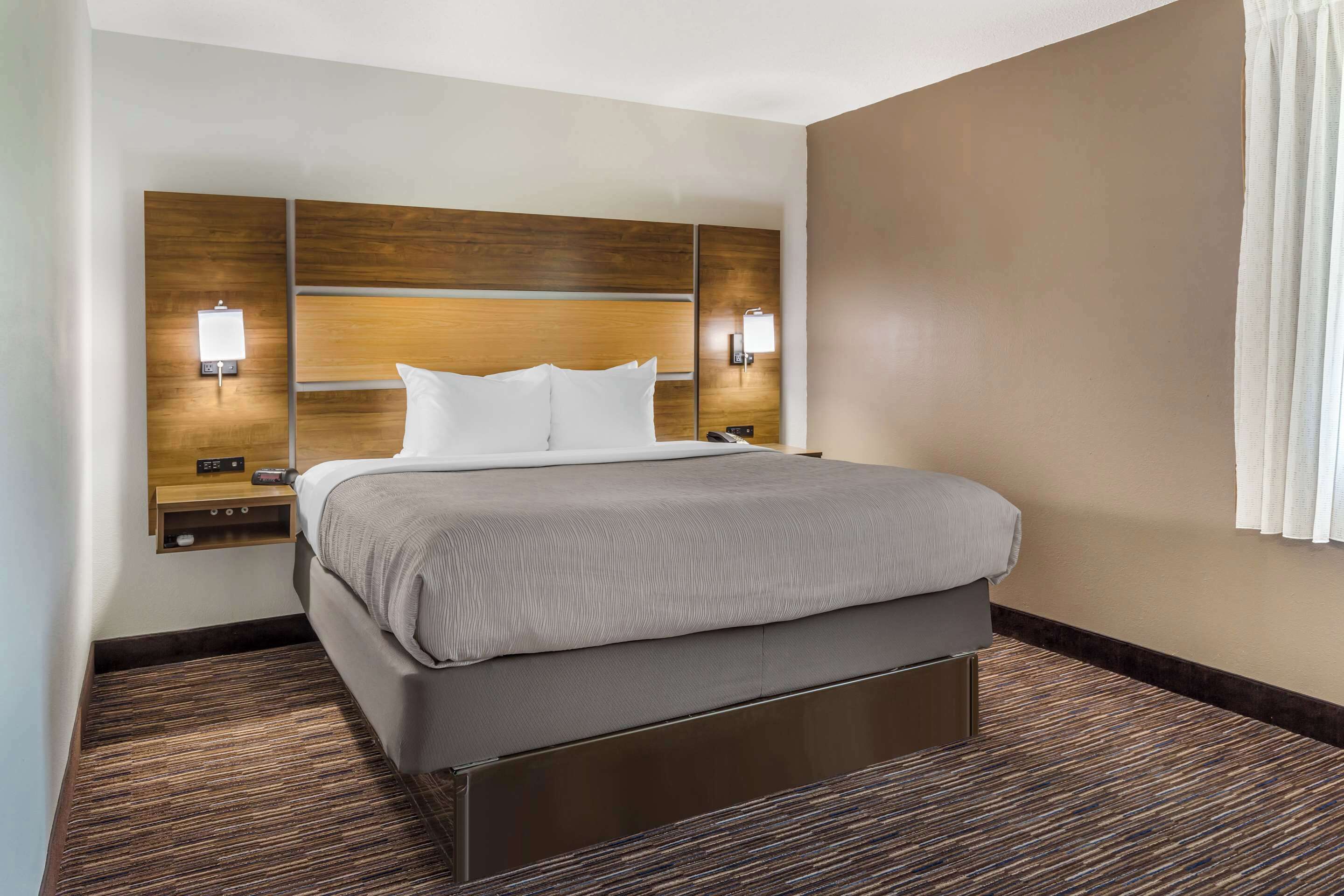 A hotel room with a large bed, wooden headboard, two bedside lamps, a nightstand, and a carpeted floor.