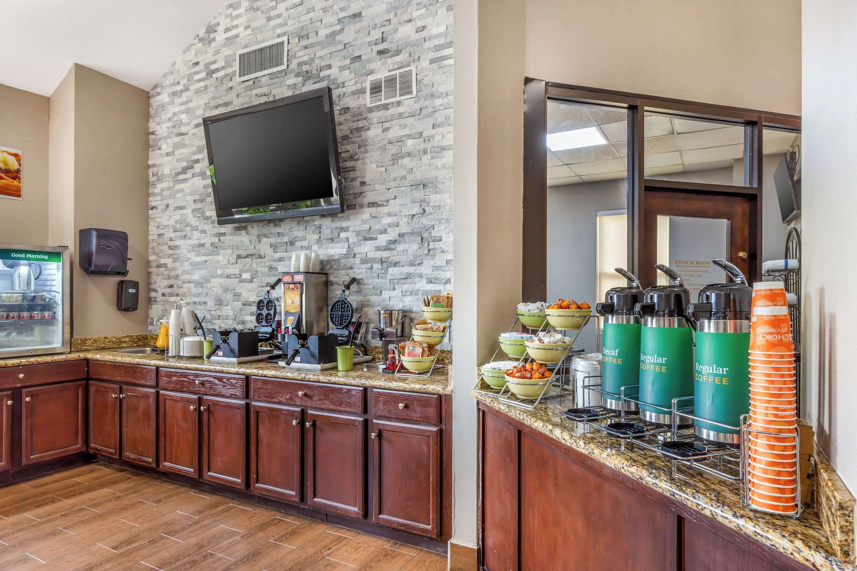 A hotel kitchen with a granite countertop, wooden cabinets, a flat-screen TV, a waffle maker, and coffee dispensers.