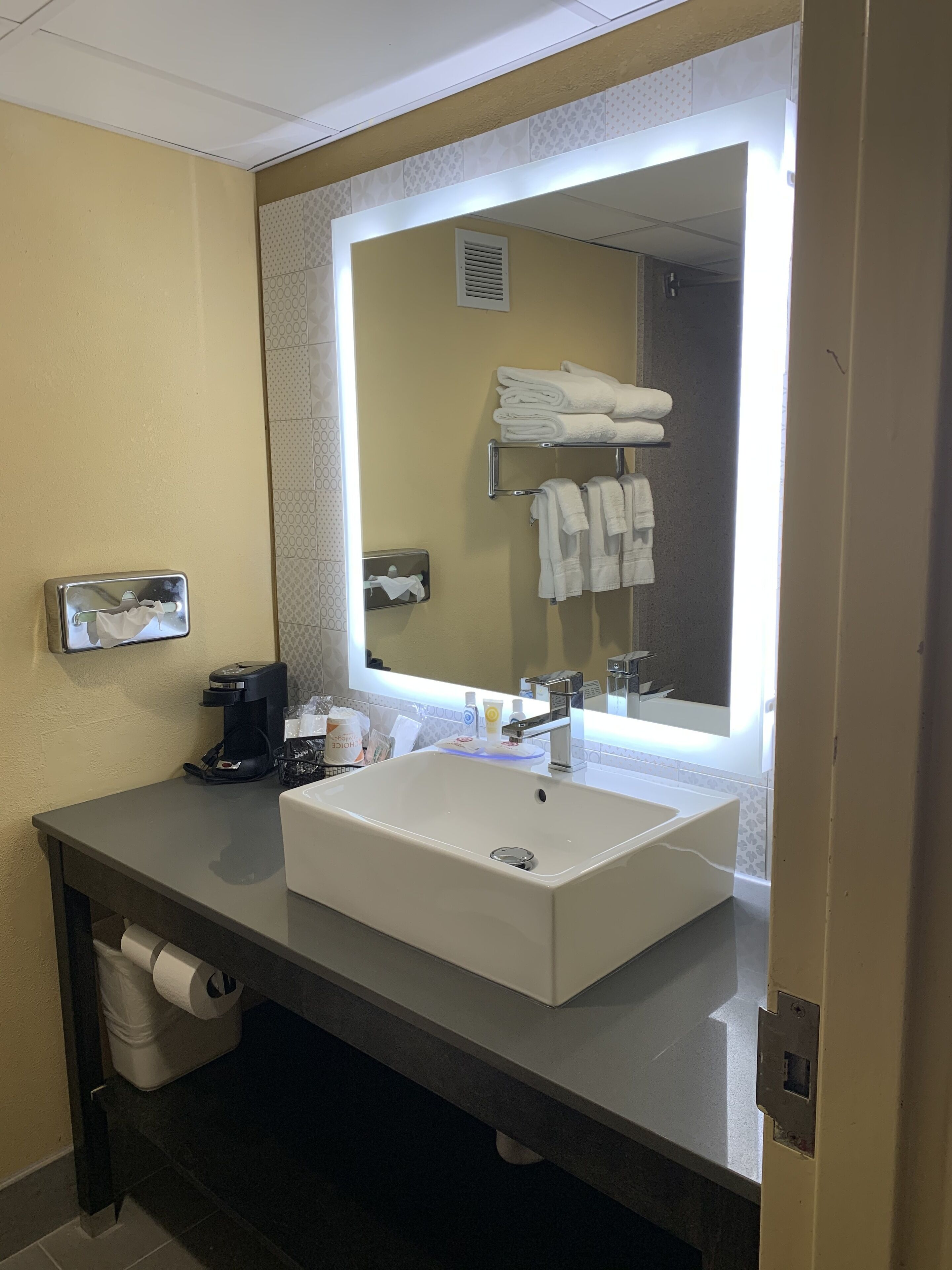 Un baño de hotel con un espejo grande, un lavabo blanco, un perchero con toallas dobladas, una cafetera y un dispensador de papel higiénico.