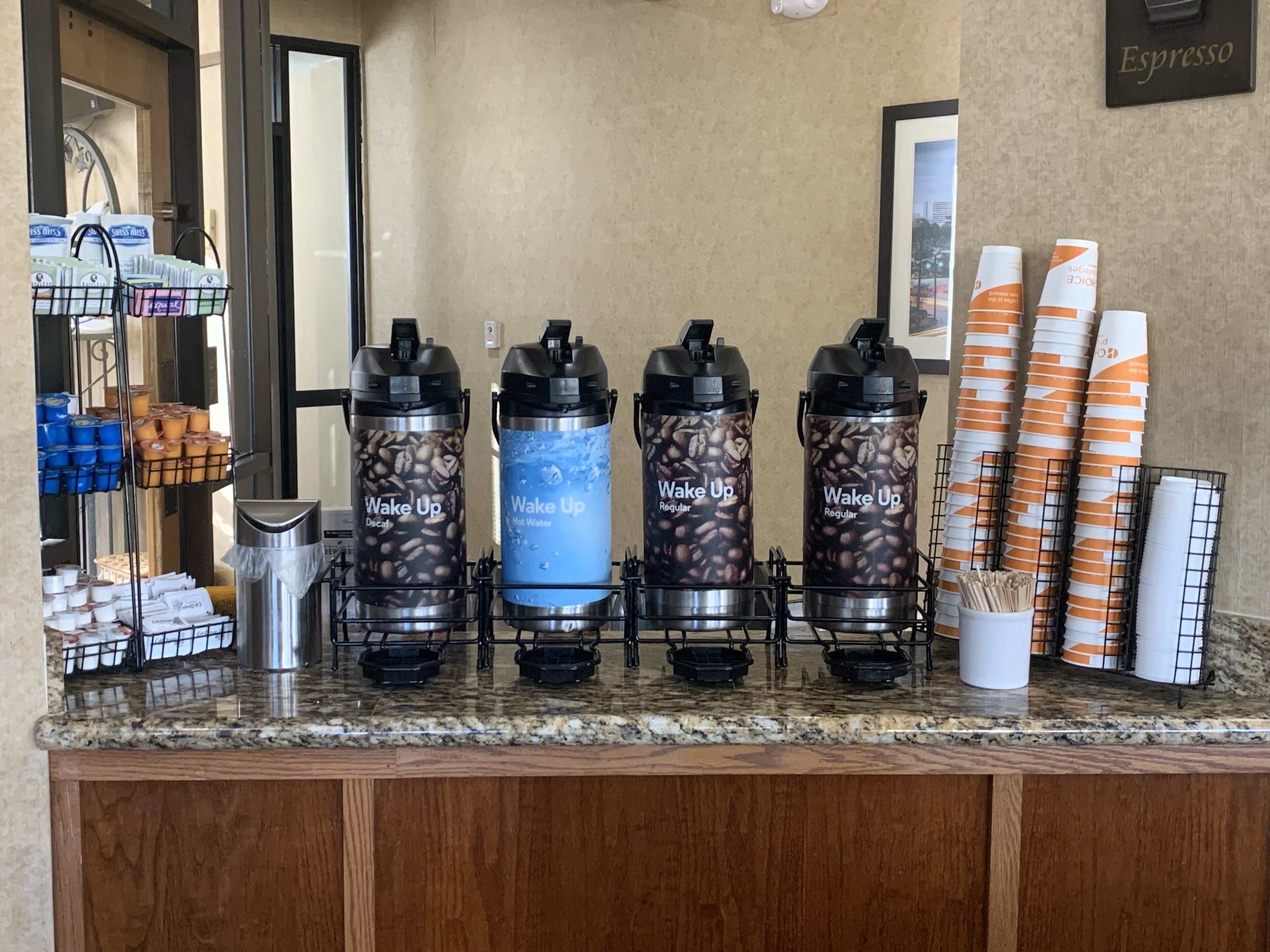 Una estación de café con cuatro dispensadores etiquetados como “Wake Up” y una variedad de cápsulas de café y vasos de papel.
