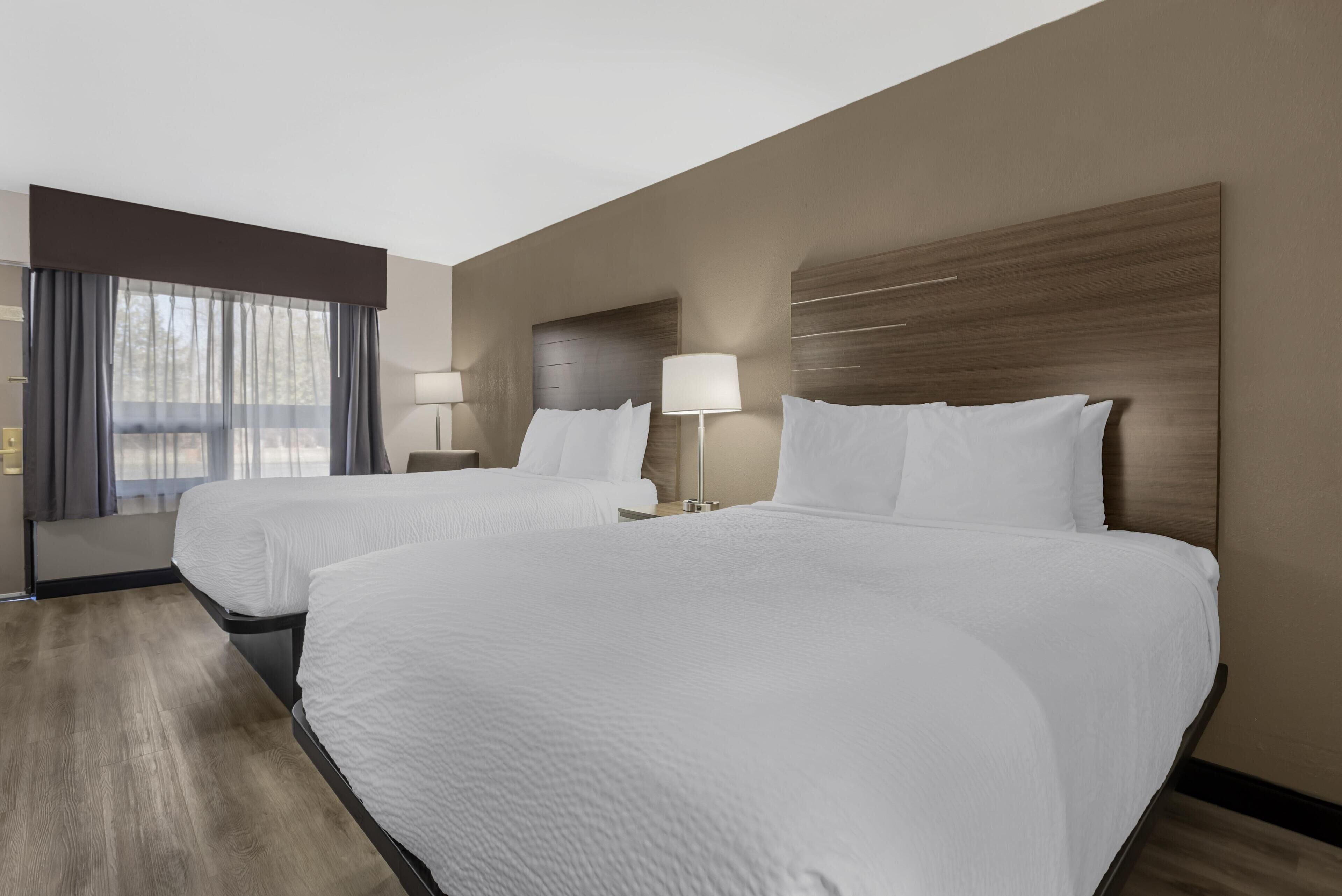Quarto de hotel com duas camas, uma janela com cortinas, uma mesa de cabeceira com abajur e cabeceira de madeira.